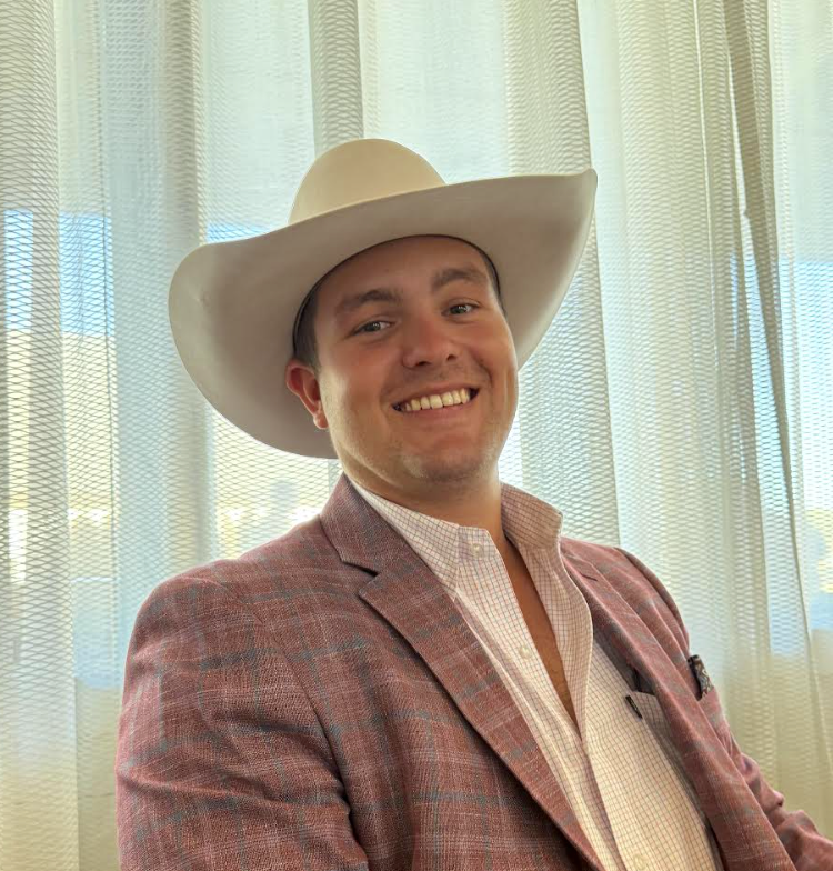 A man smiling, wearing a white cowboy hat, a checkered dress shirt, and a pinkish blazer, sitting in front of light-colored curtains.