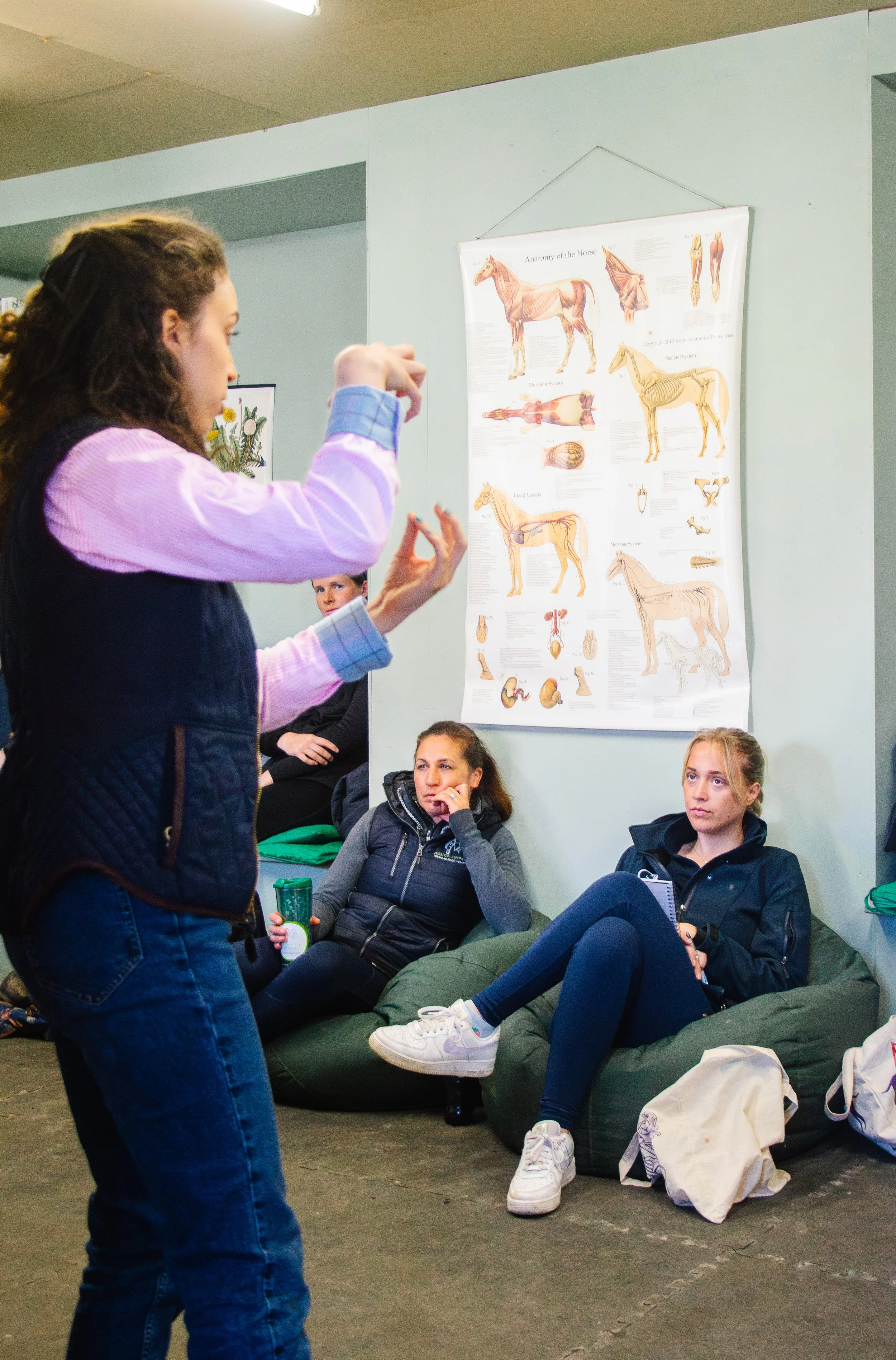 A woman standing and talking to a group of seated young women in a room with educational posters about the horse's anatomy.