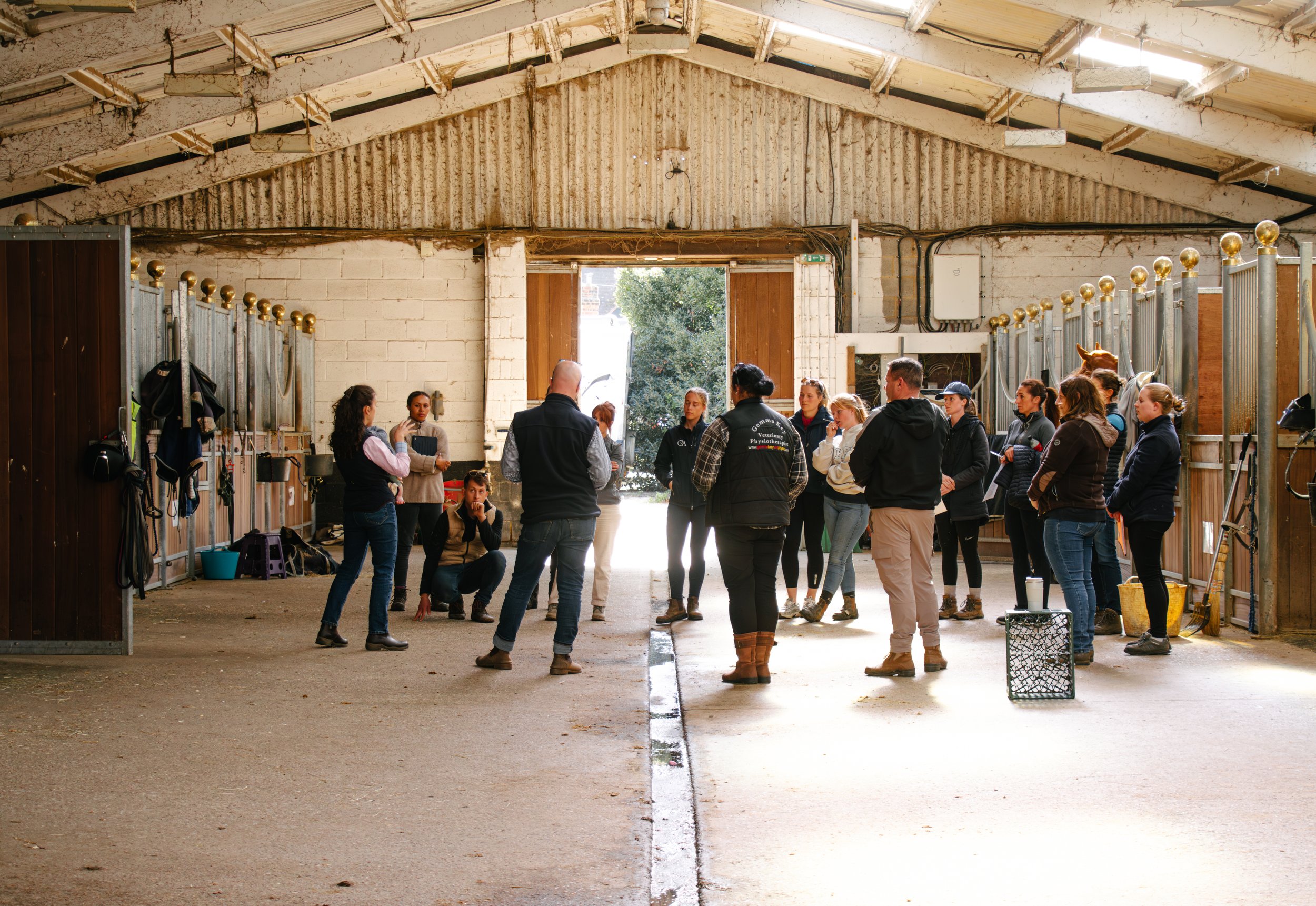 A group of people gathered inside a horse stable, with horse stalls on either side. The open barn door reveals trees outside.