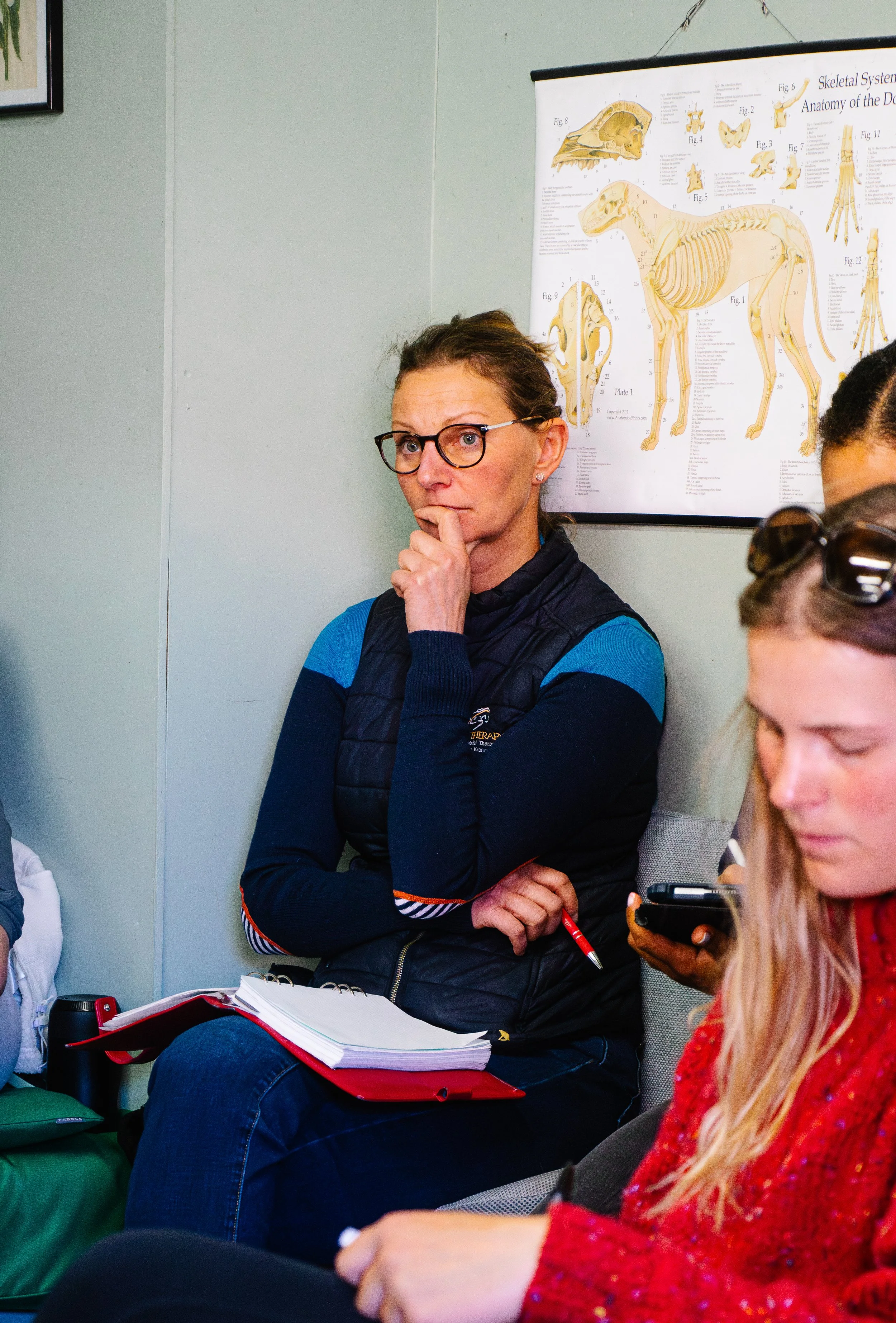 A woman with glasses and a black vest sitting in a meeting room, holding a red pen and notepad, with a puzzled expression. Behind her, a skeletal system diagram is on the wall, and another woman in a red sweater is using a smartphone.