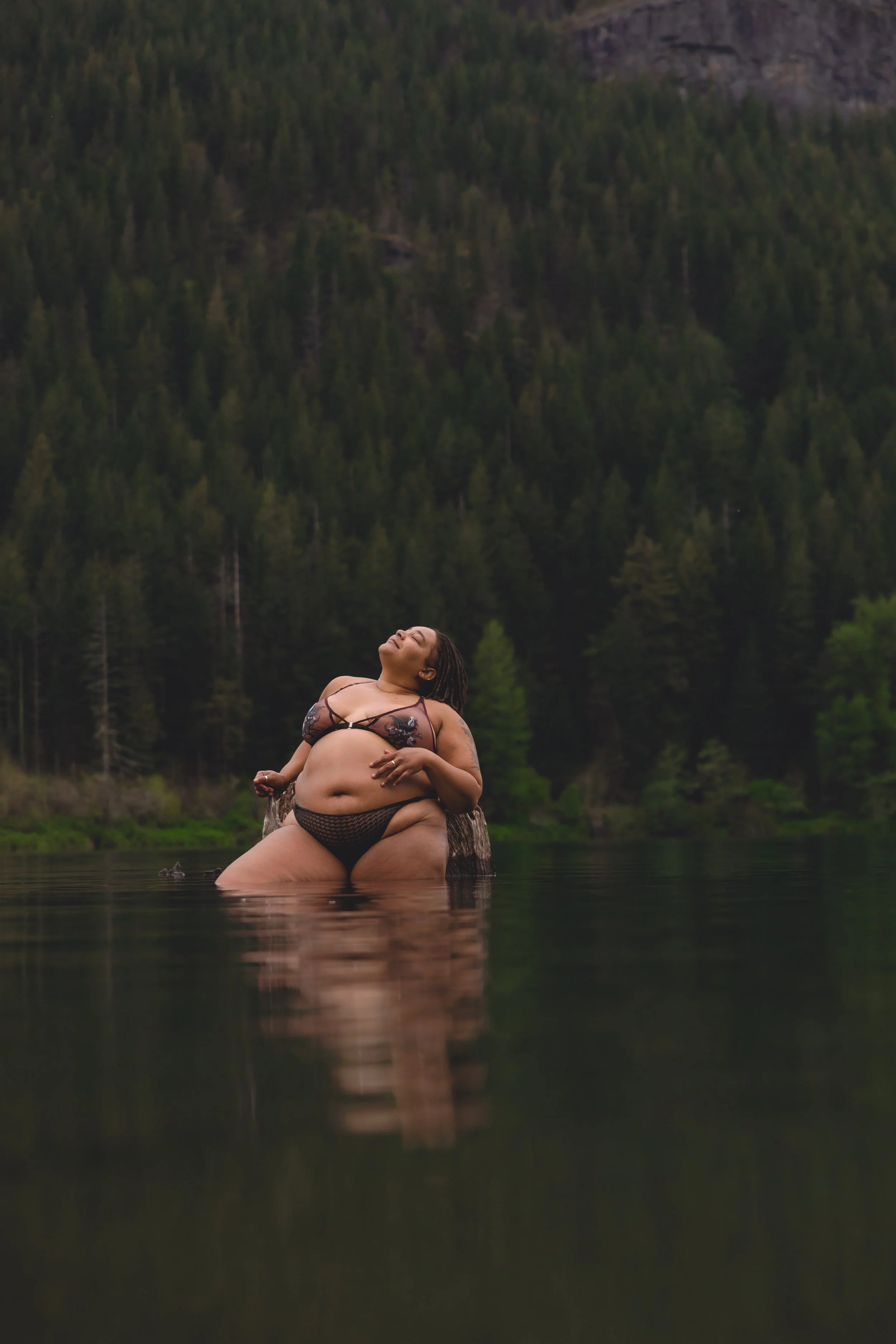 Woman standing in a body of water with a forested mountain in the background, wearing black lingerie and smiling.