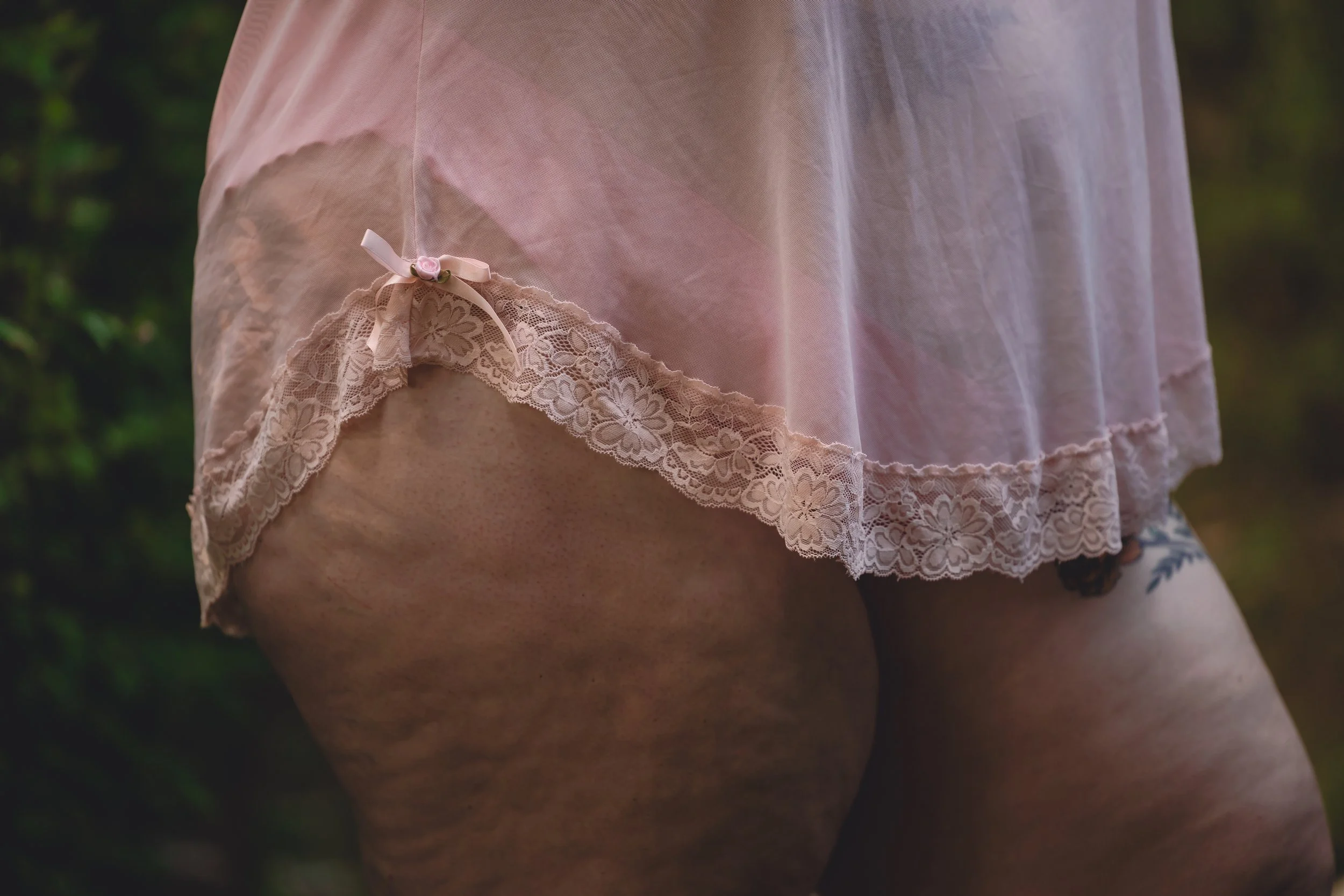 Close-up of a person's upper thigh and hip, wearing a sheer pink lace slip with lace trim and a small pink bow, outdoors with blurred greenery in the background.