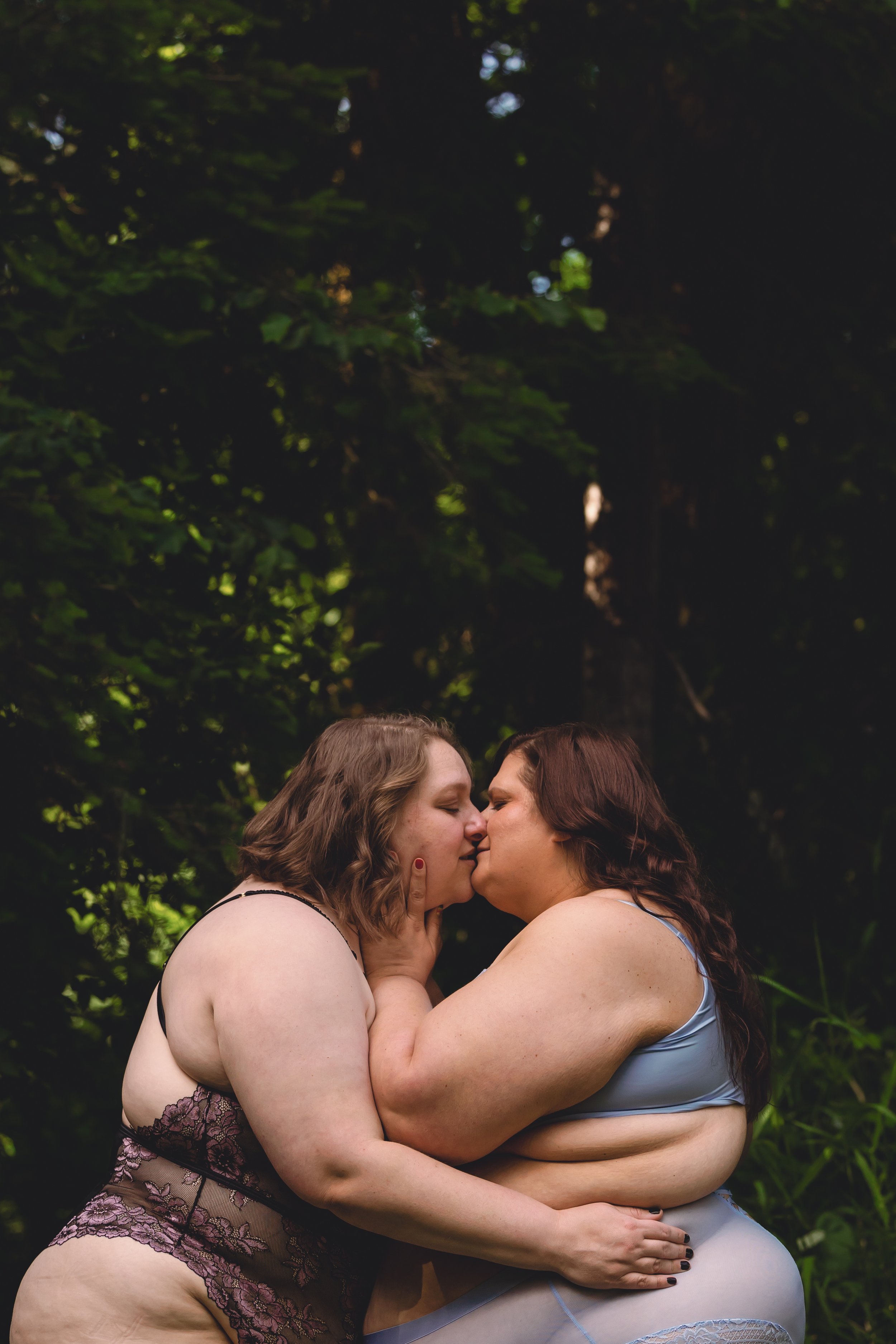 Two women are kissing in a wooded outdoor setting.