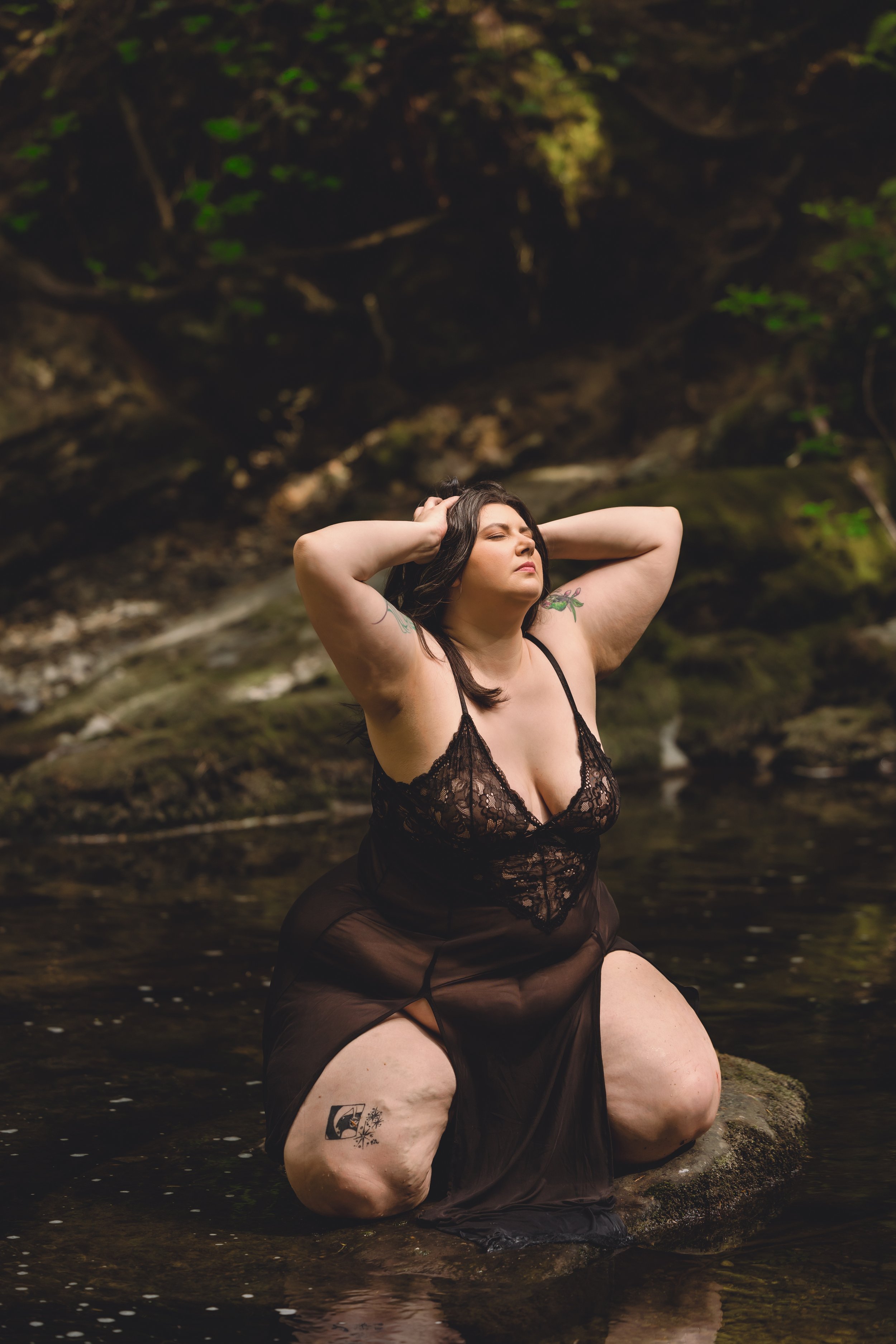 A woman with dark hair and tattoos on her arms and thigh kneeling in a shallow creek, wearing black lace and sheer black dress, with her hands behind her head and eyes closed, surrounded by a natural forest setting.