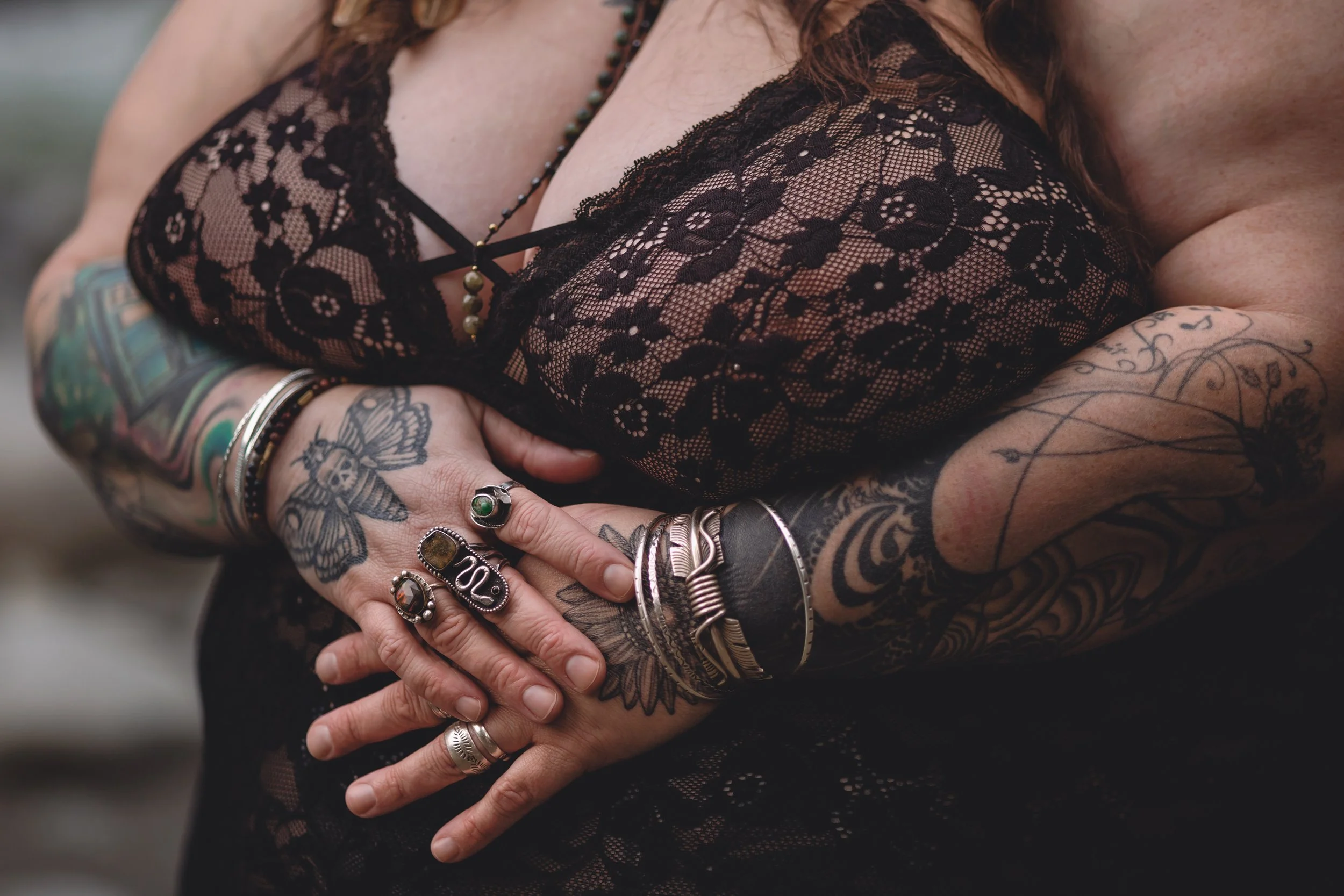Close-up of a woman's arms crossed over her chest, showing tattoos and multiple rings, wearing a black lace top.