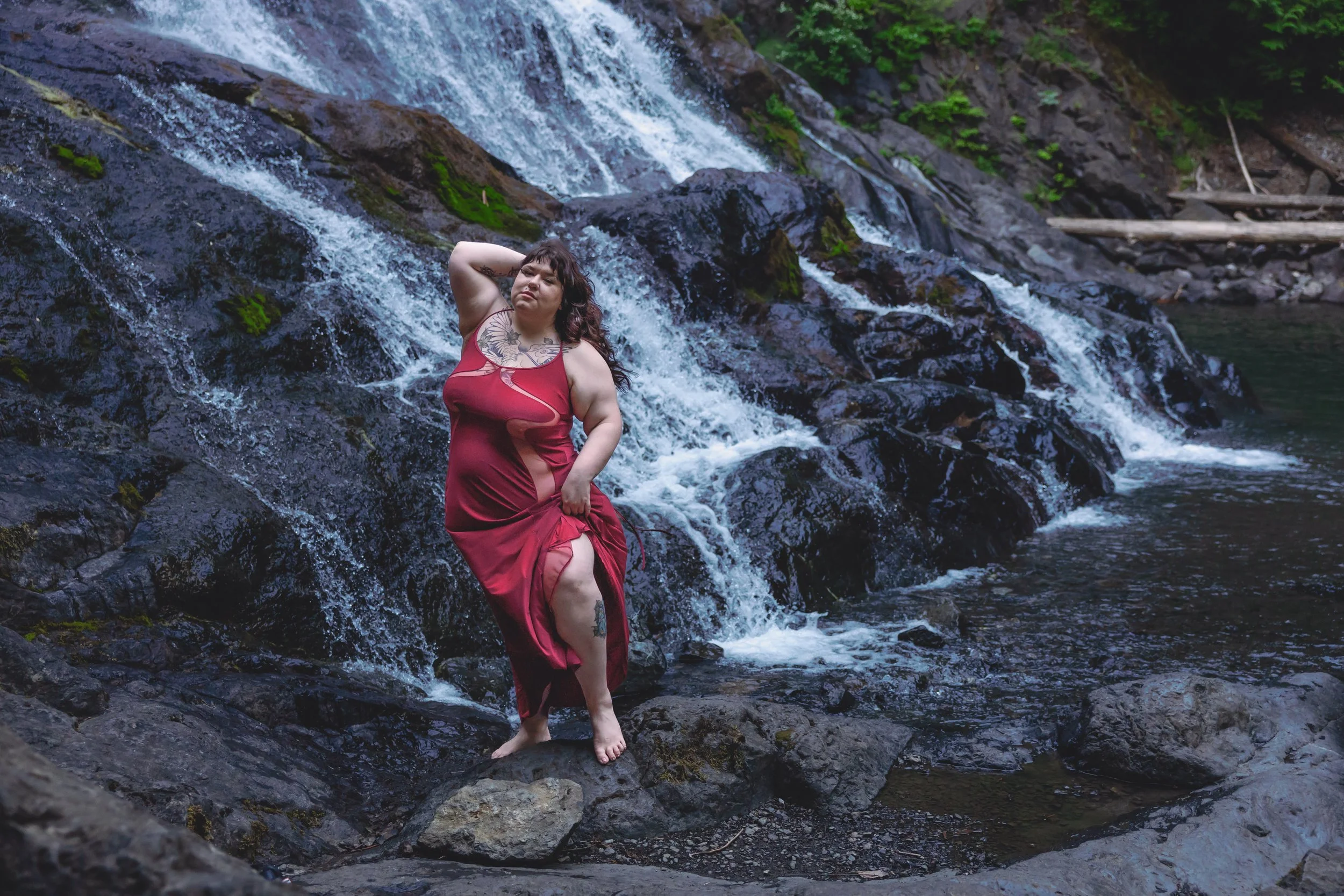 A woman standing barefoot on rocks in front of a cascading waterfall, wearing a red dress with a slit, posing with one hand behind her head and gazing into the distance.