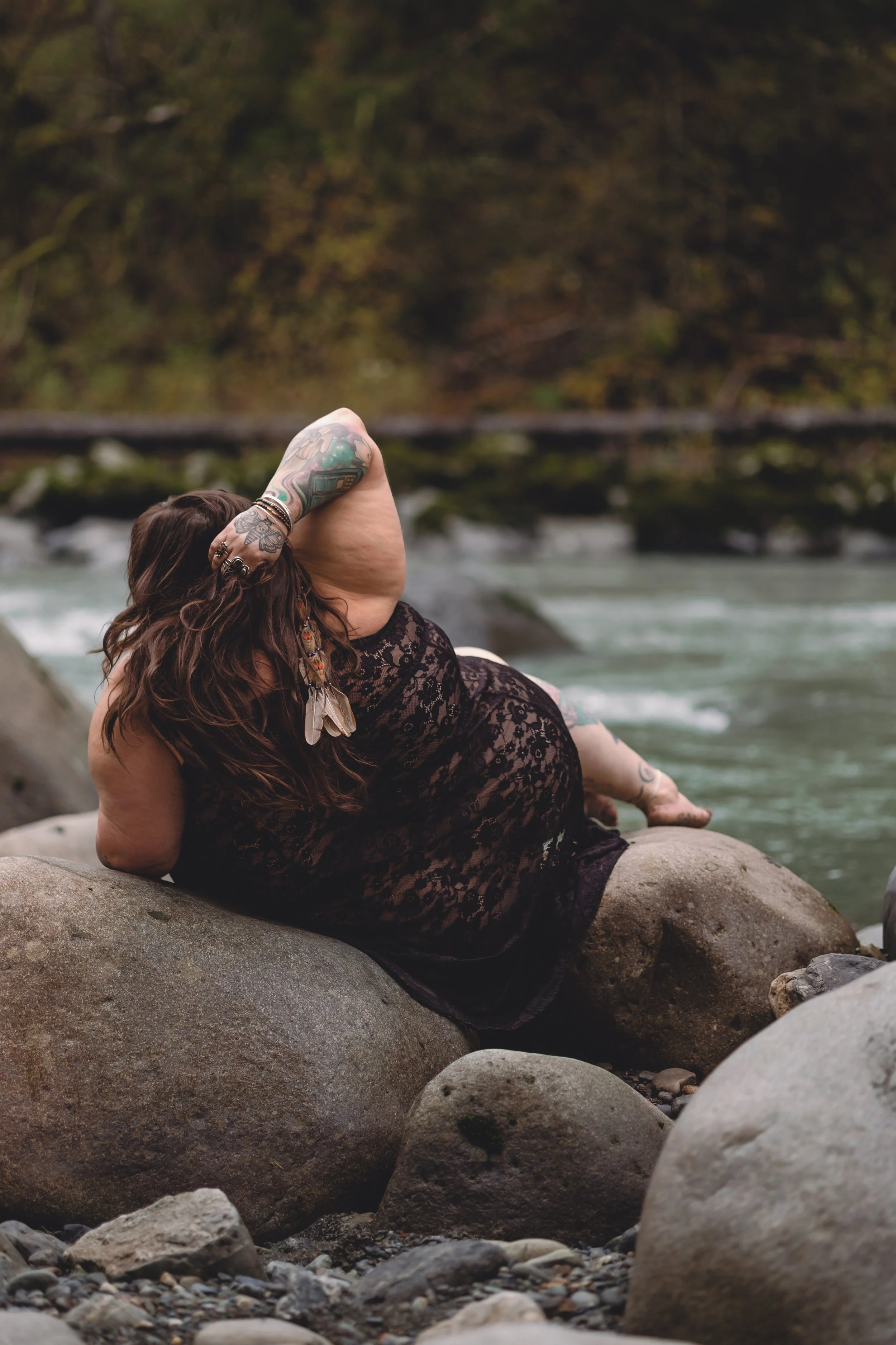 A woman with long dark hair, tattoos, wearing a black lace dress, reclining on large rocks by a river, with a blurred forest background.
