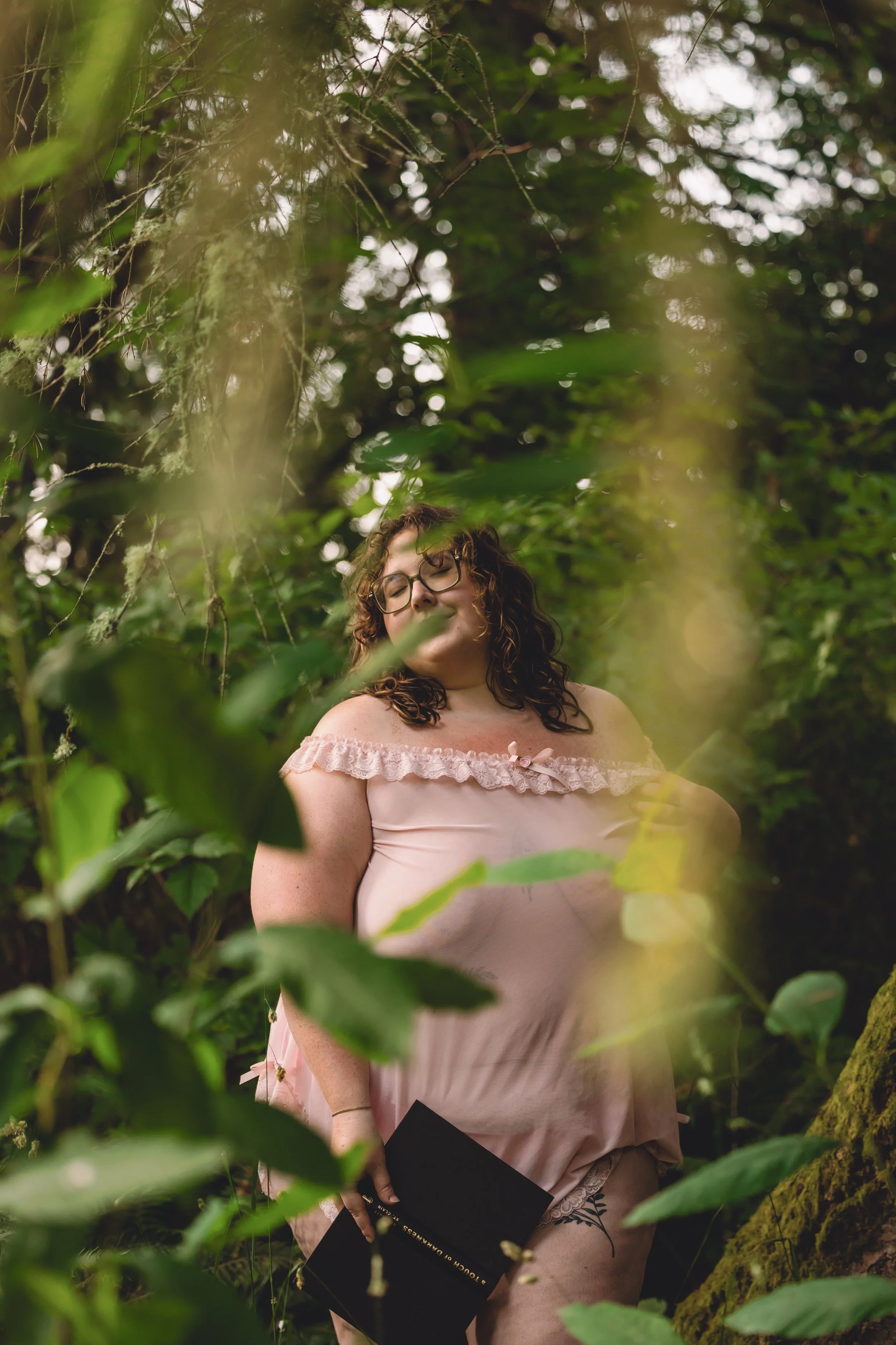 A woman with curly hair and glasses wearing a pink, off-shoulder dress standing in a lush green forest, smiling with eyes closed while holding a black graduation diploma.