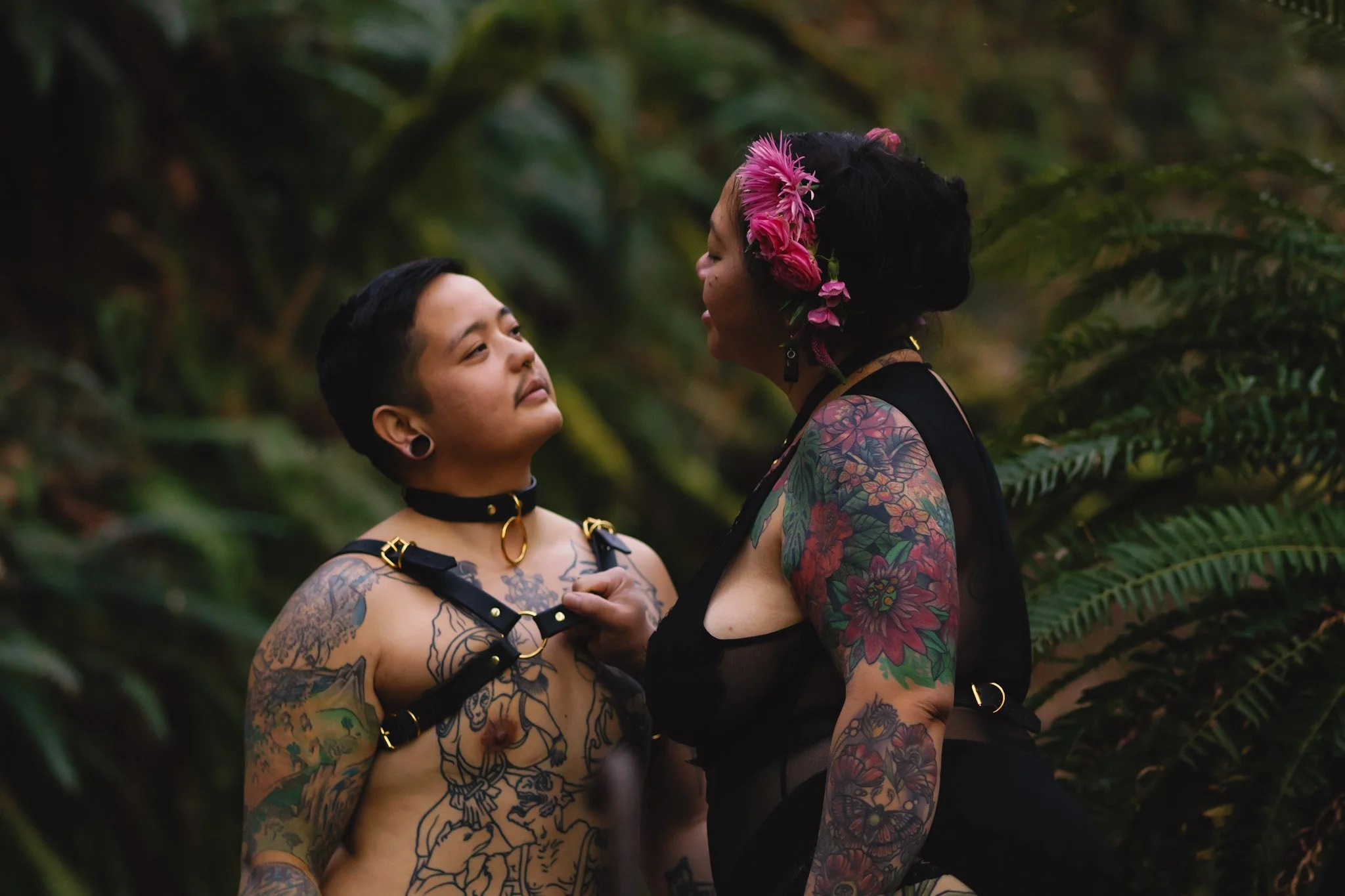 Two tattooed women in black outfits with harnesses standing close together in a lush green jungle, one woman has pink flowers in her hair.