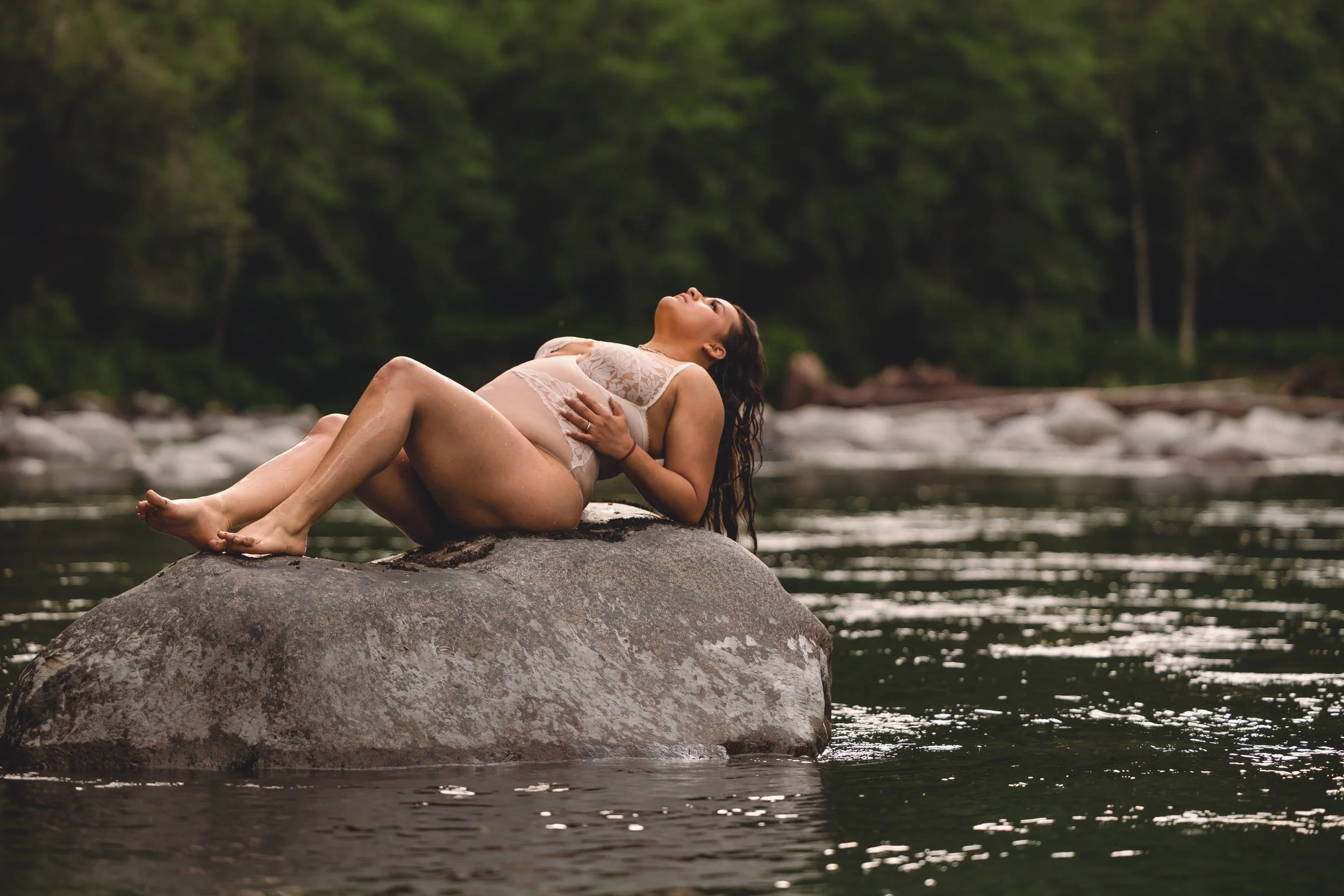 A woman lying on a large rock in a river, wearing lingerie, with her head tilted back and eyes closed, surrounded by lush green trees.