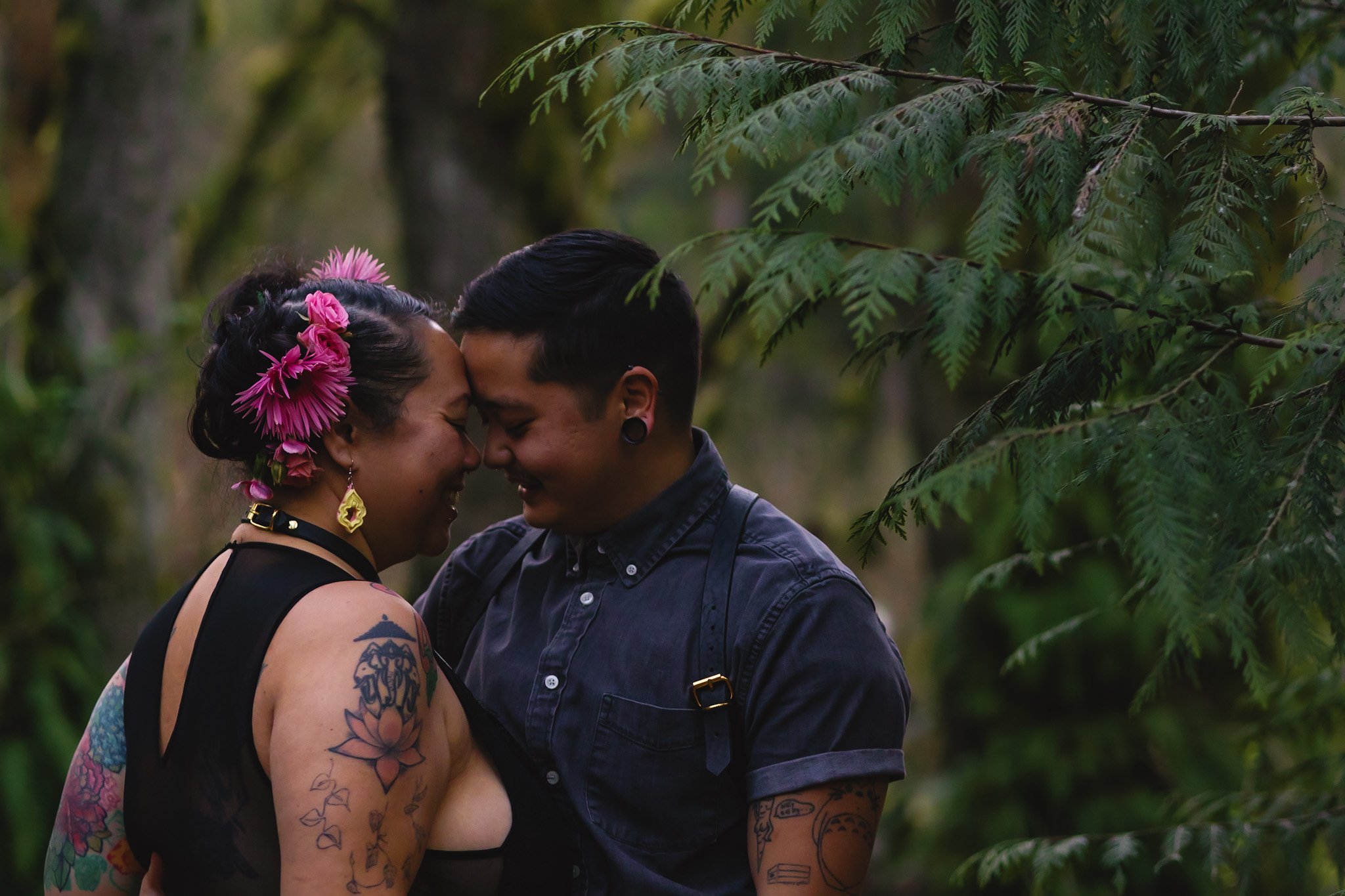 A couple shares an intimate moment, touching foreheads and smiling, surrounded by green foliage in a forest.