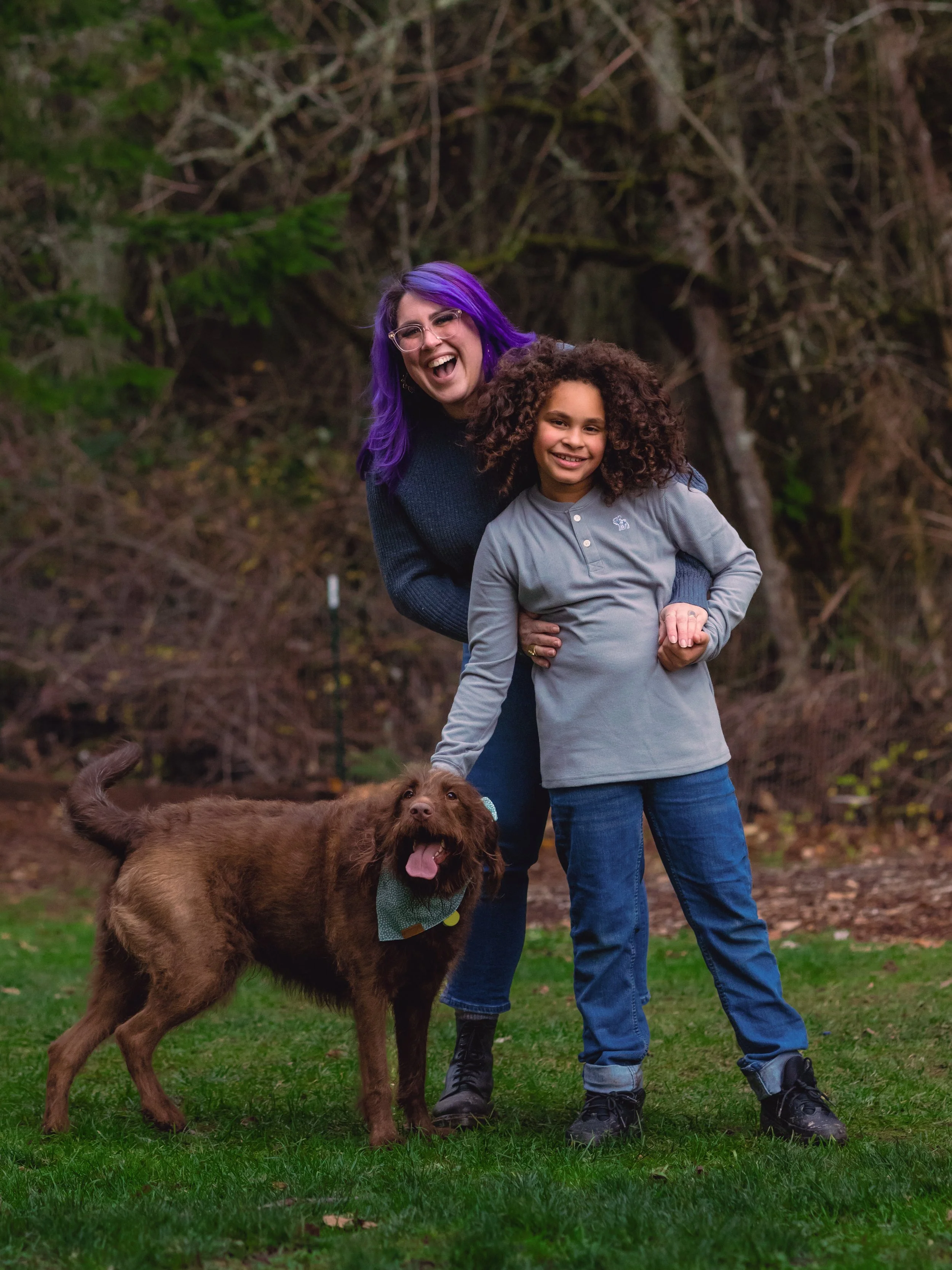 A woman with purple hair, a girl with curly hair, and a brown dog are outdoors on a grassy area with trees in the background, smiling and enjoying themselves.