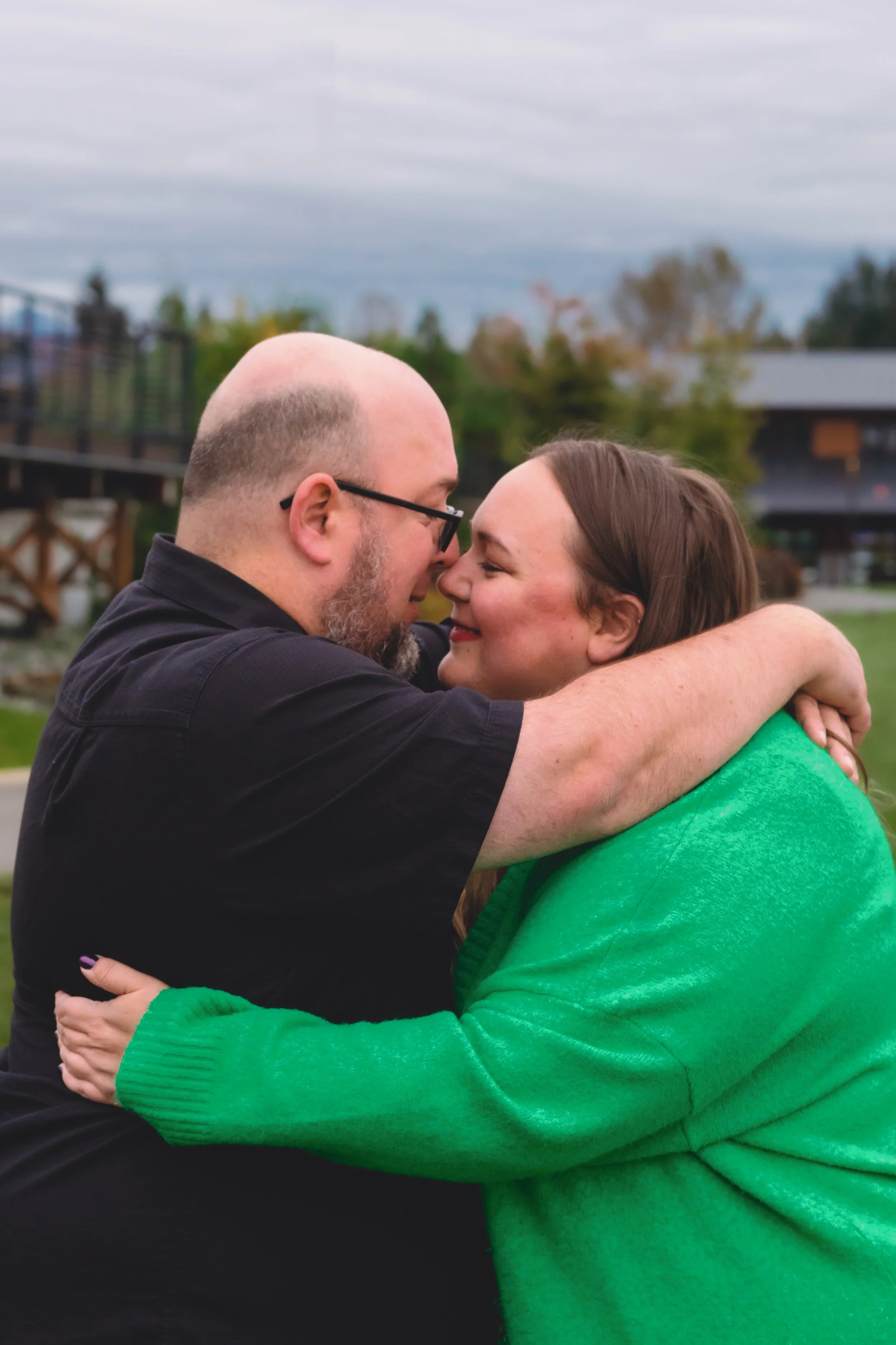 A man and a woman embracing outdoors, their faces close together, smiling with closed eyes, against a cloudy sky and a blurry background of trees and buildings.