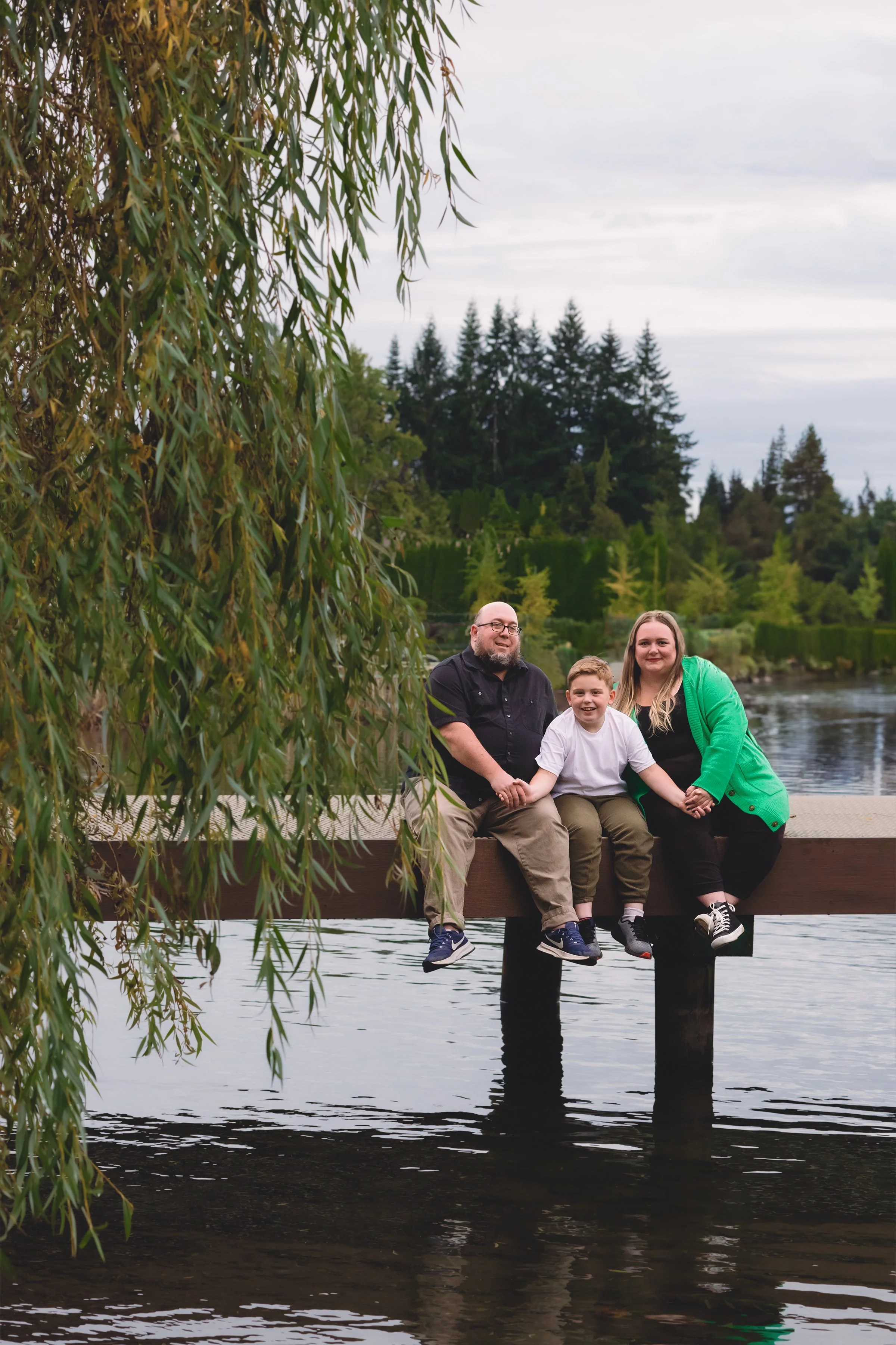 A family of three sitting on a wooden dock over a lake, holding hands and smiling, with trees and a cloudy sky in the background.