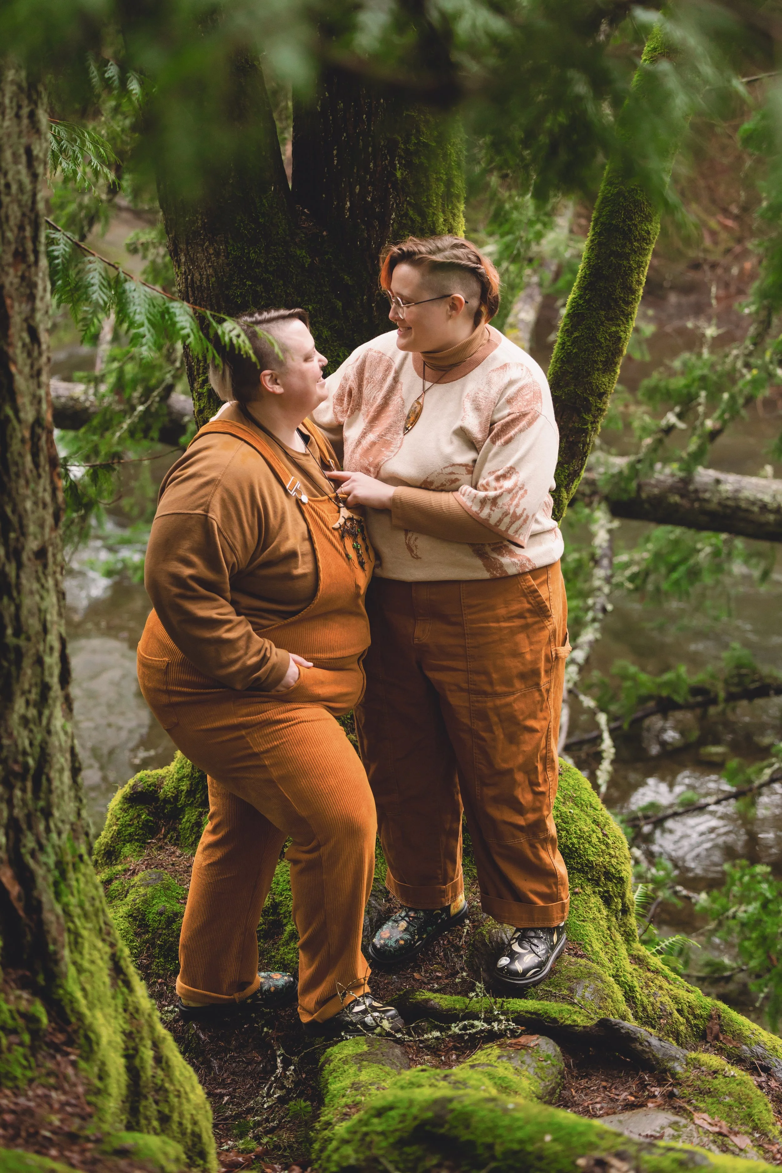 Two people standing close together in a mossy forest, smiling at each other. One has short hair and glasses, the other has longer hair in braids. Both are dressed in earthy-toned clothing, standing on a moss-covered log.