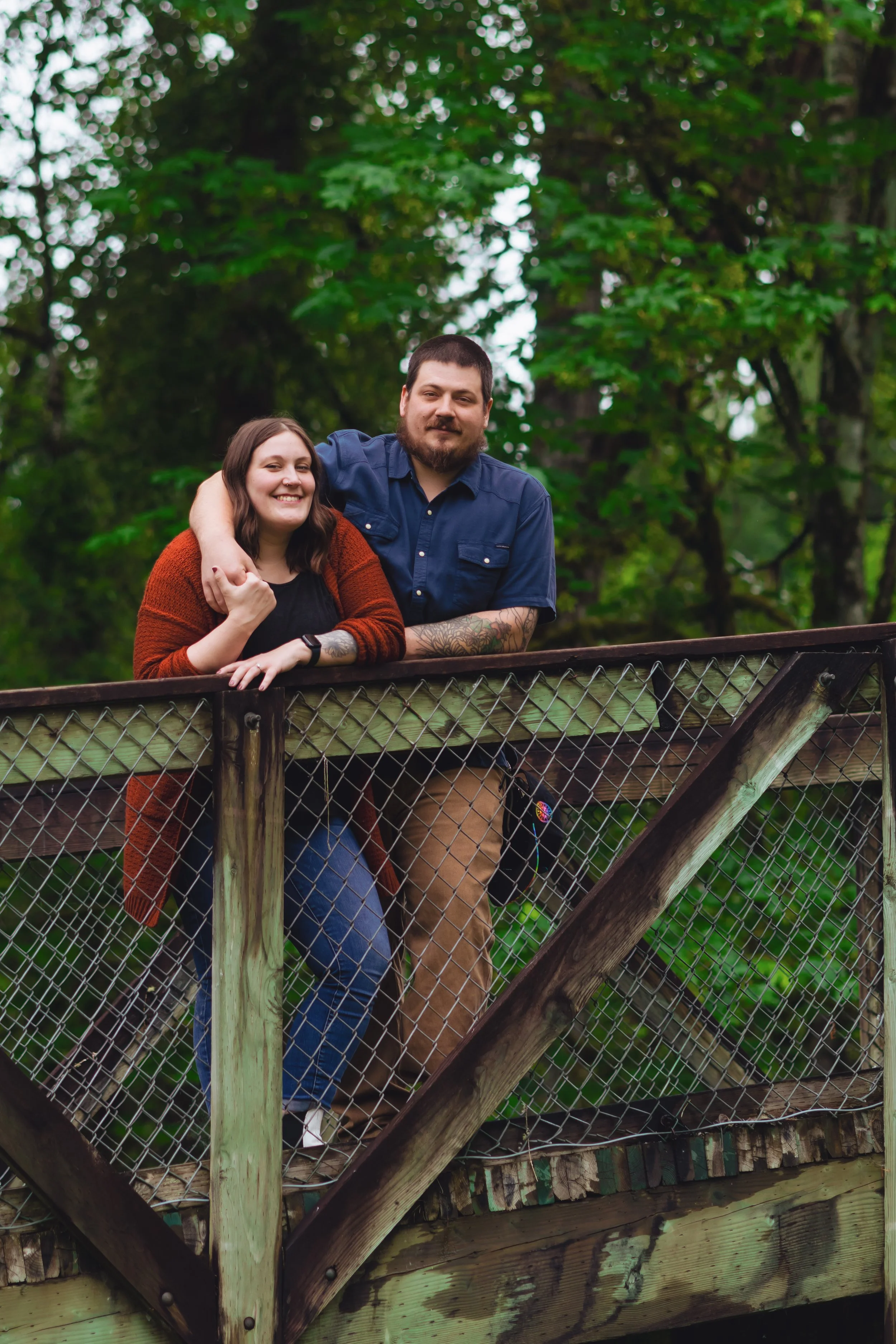A smiling couple leaning on a wooden fence on a bridge surrounded by lush green trees.