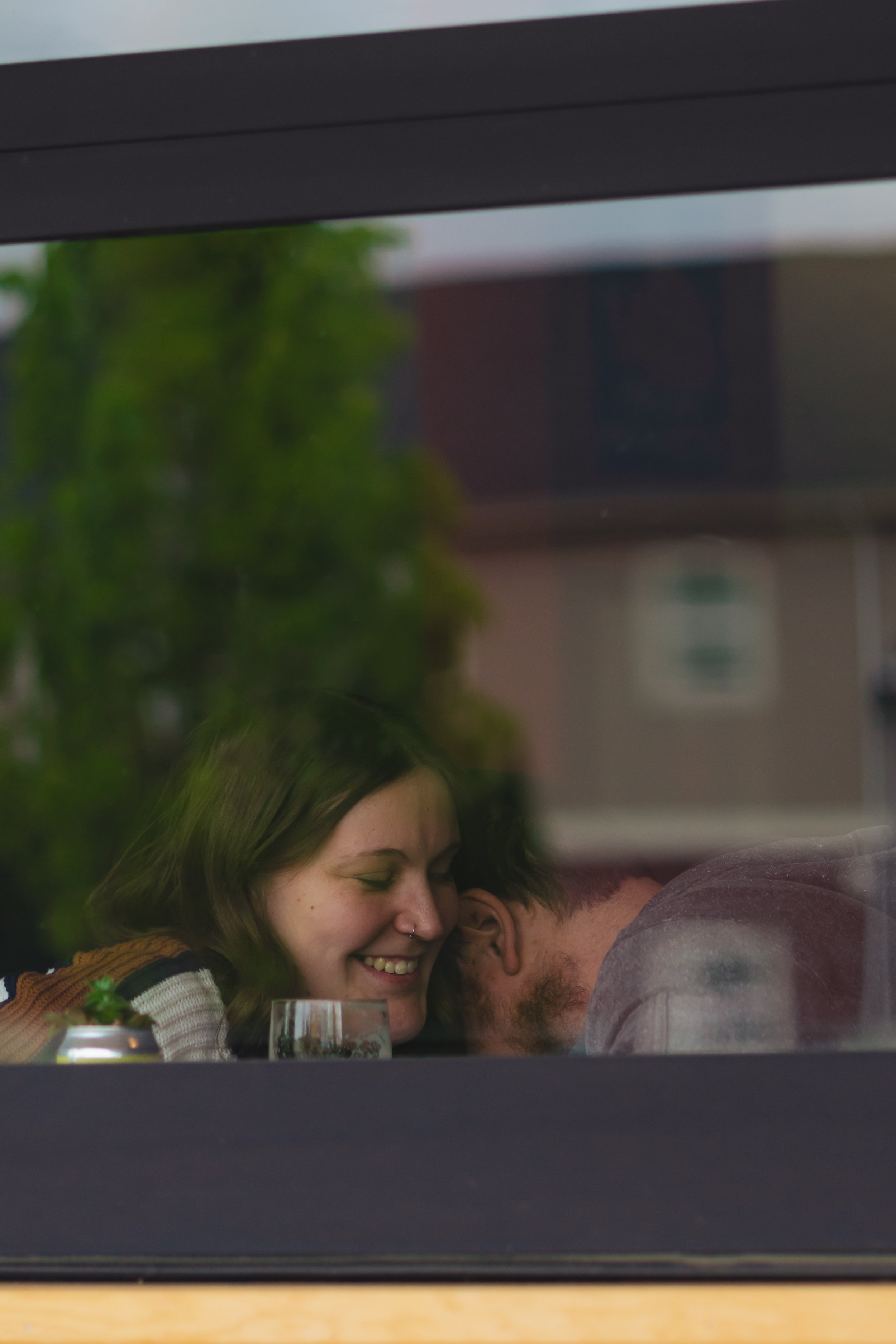 Two people smiling and leaning their foreheads together, viewed through a window with reflections of trees and buildings.