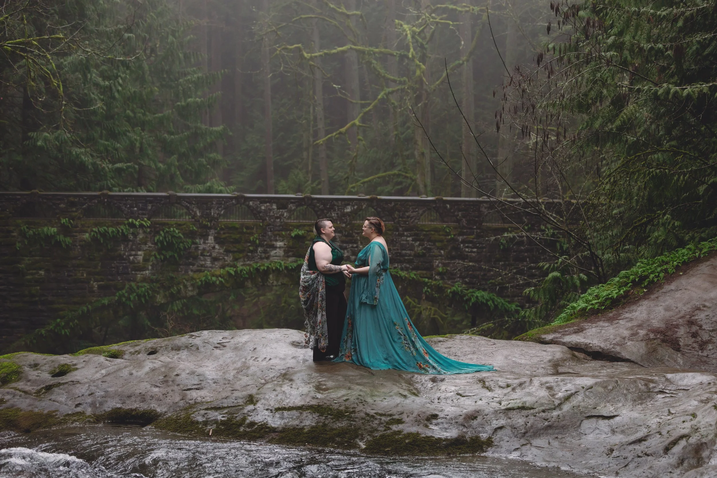 Two women holding hands and looking at each other on a large rock near a stream in a forest. One woman wears a teal dress with floral embroidery, and the other wears a dark outfit with a patterned shawl. A bridge is visible in the background amid tal