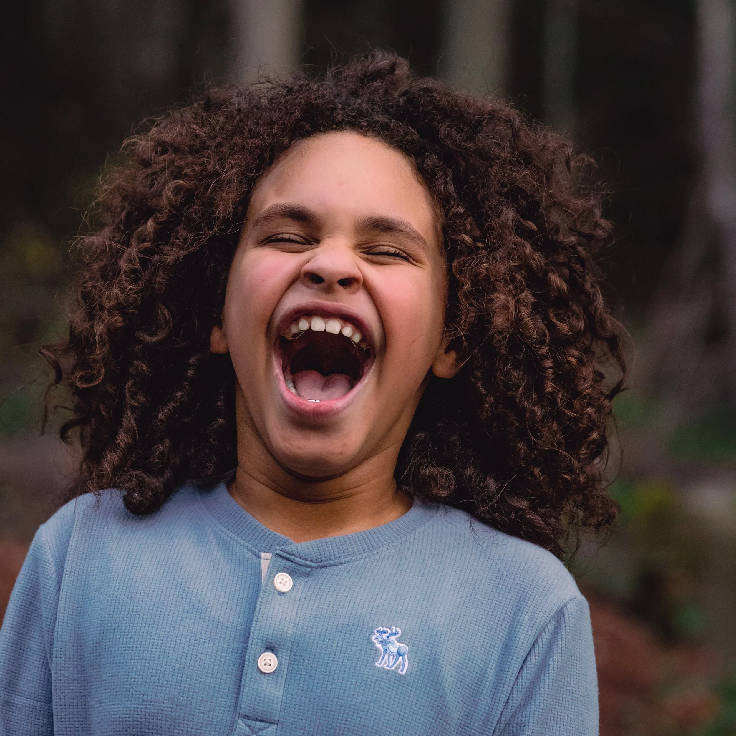 A young girl with curly hair laughing outdoors, wearing a light blue shirt.