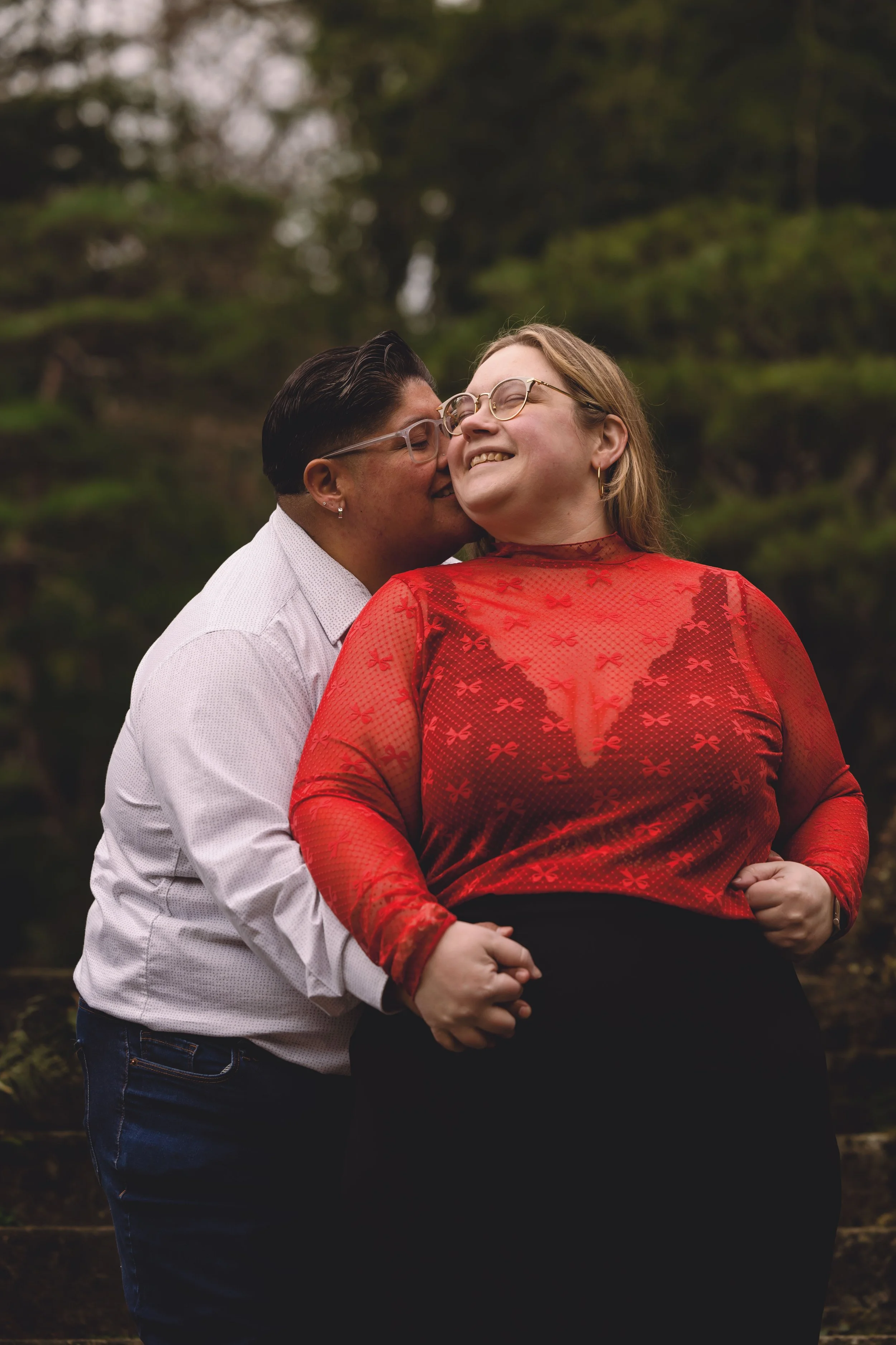 A diverse couple in love outdoors, with the woman smiling happily as her partner kisses her cheek.