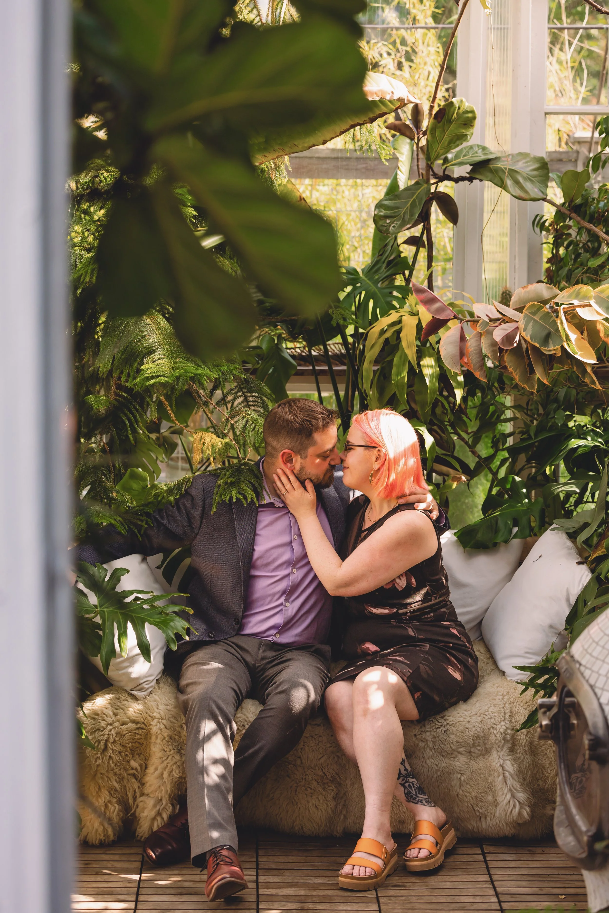 A couple is sitting closely together in a lush, greenhouse-like setting surrounded by large green plants. They are touching foreheads and smiling at each other, creating a romantic atmosphere.