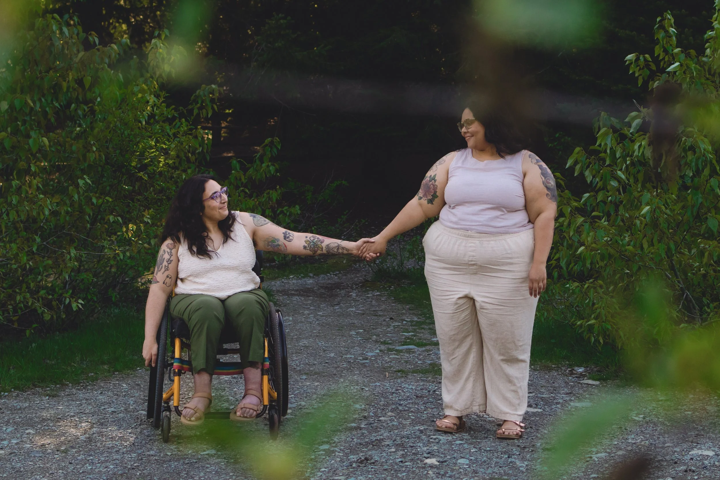 Two women, one in a wheelchair and one standing, holding hands and smiling at each other in a lush outdoor setting.