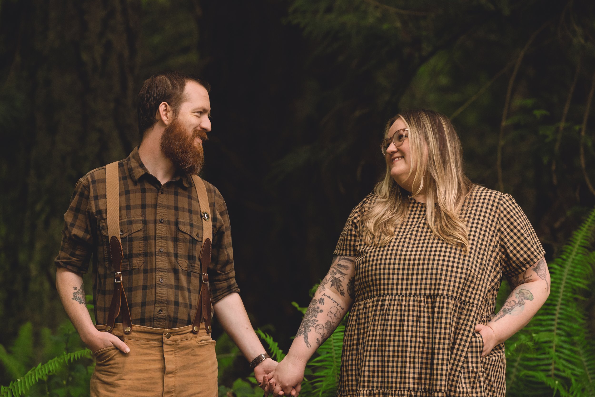 A man and woman holding hands in a forest, smiling at each other.