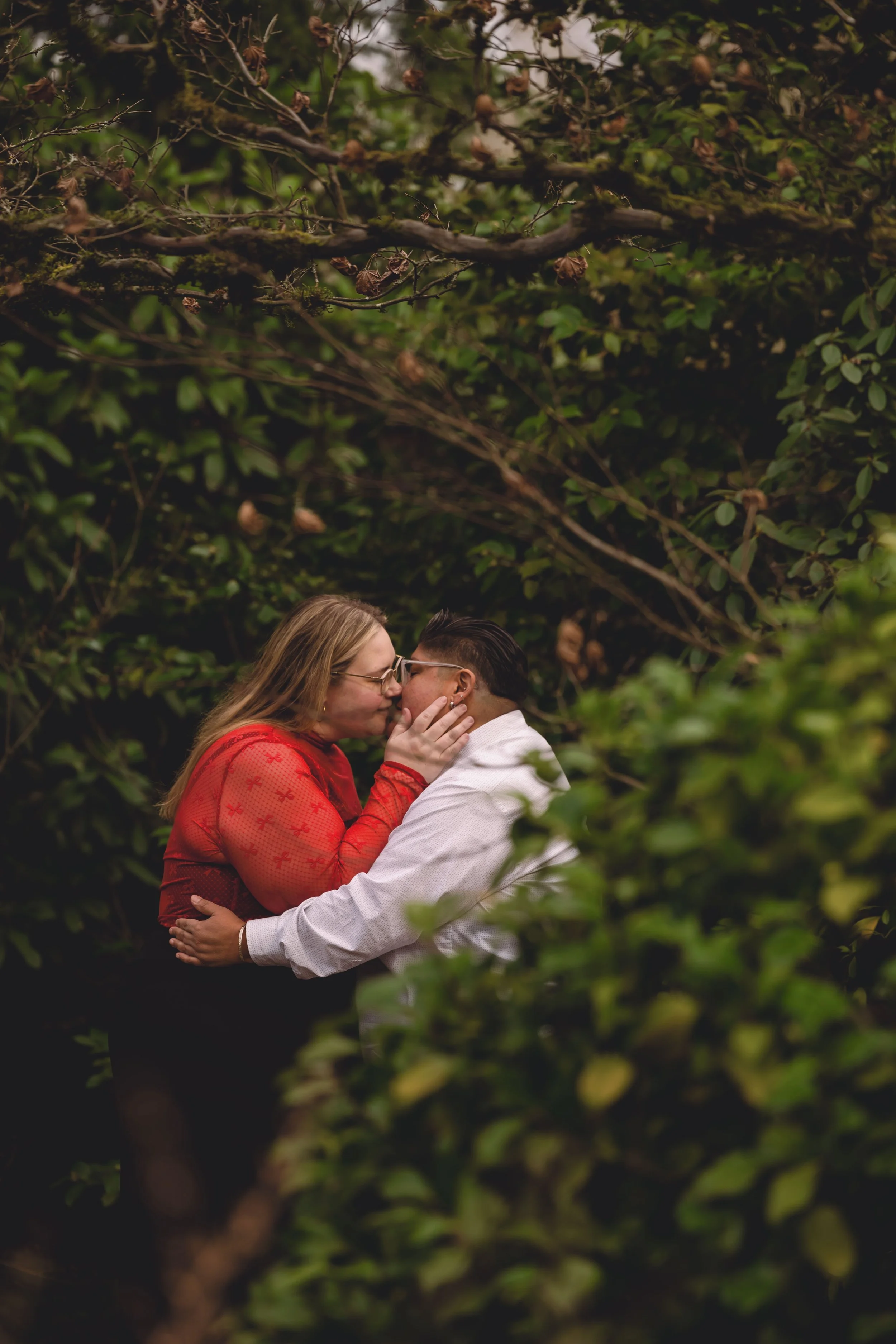 A couple sharing a kiss in a lush green, outdoor setting surrounded by dense foliage and tree branches.