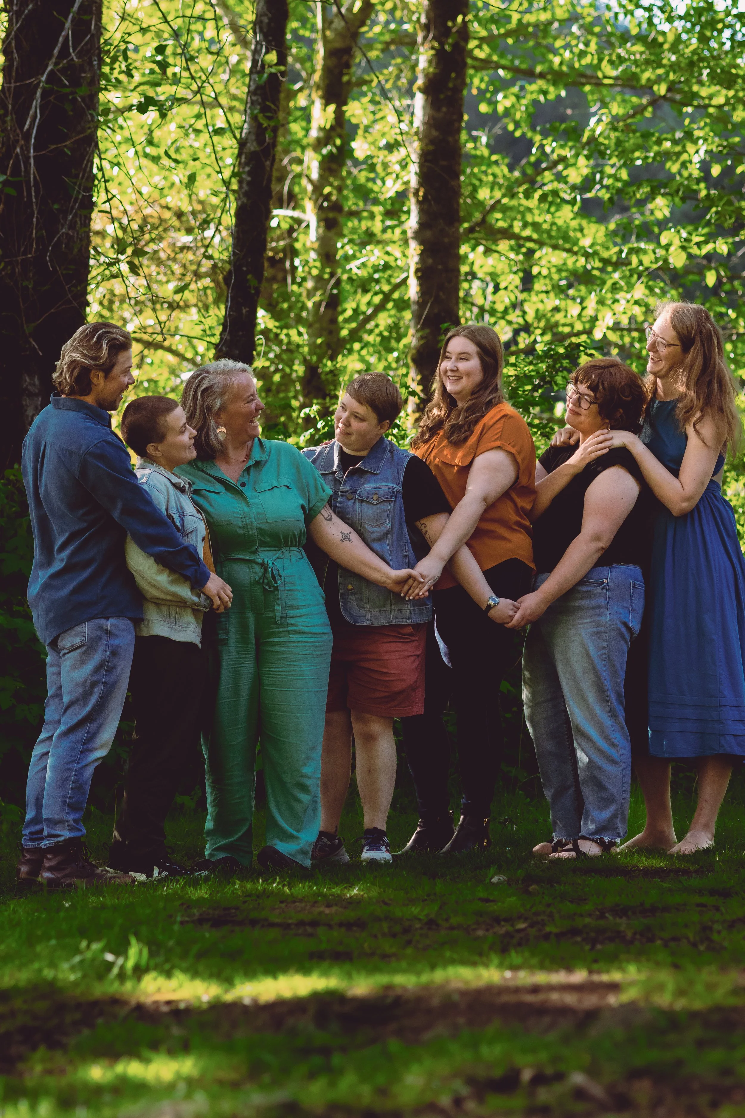 A group of seven people, including children and adults, are standing together in a forest, smiling and holding hands.
