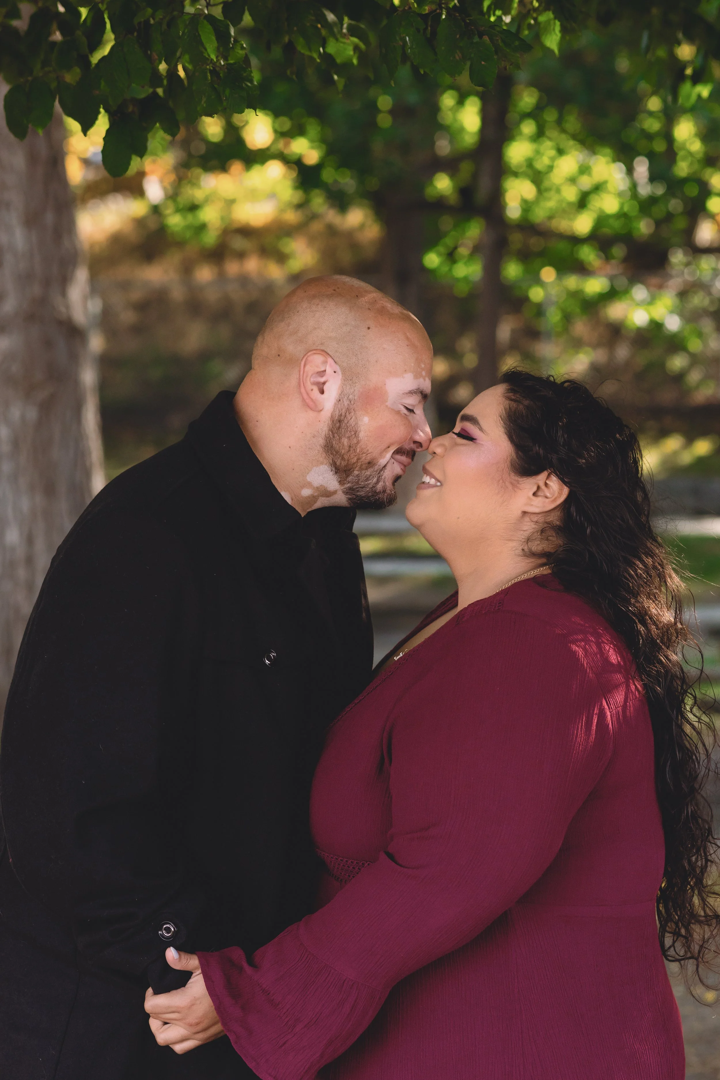 A couple standing close together outdoors, holding hands, facing each other with their noses touching, smiling, surrounded by green trees and sunlight.