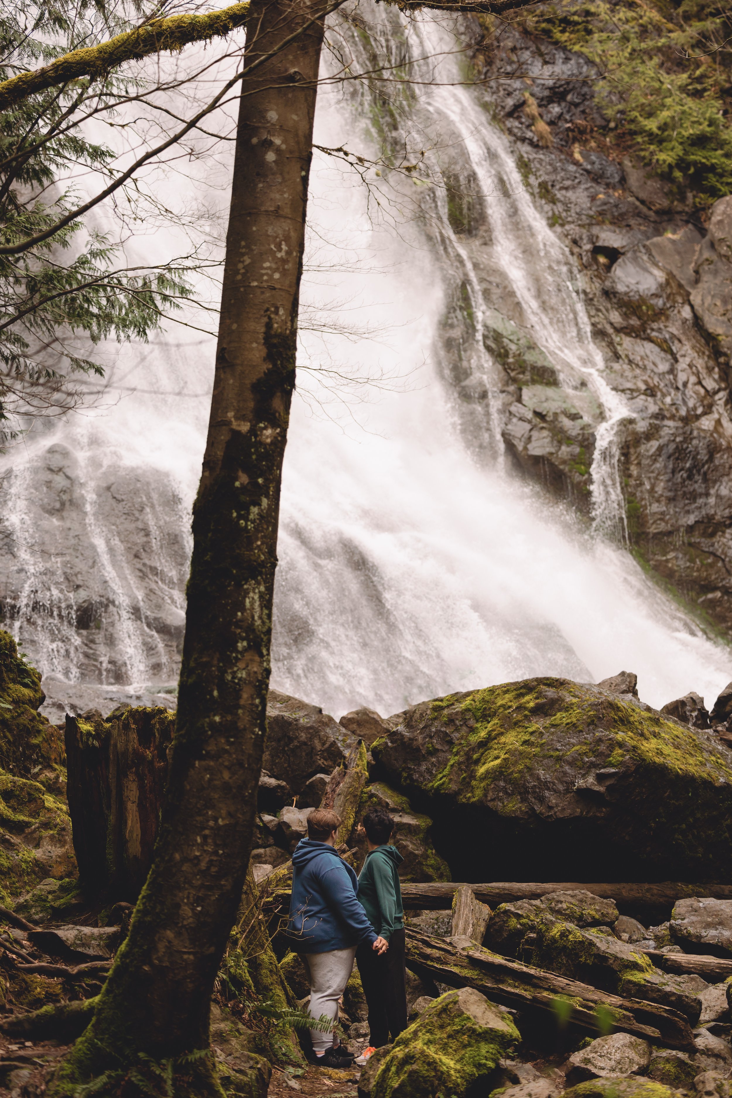 Two people holding hands and facing a large waterfall in a forested area with moss-covered rocks and trees.