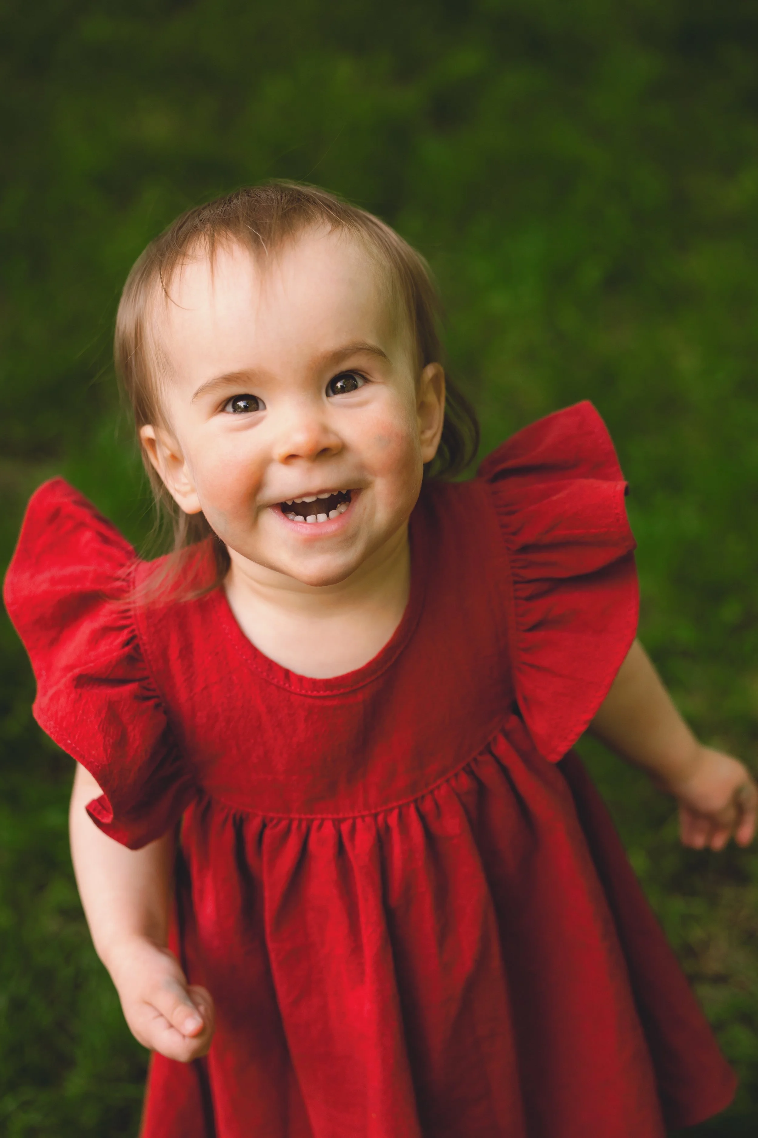A young girl in a red dress smiling outdoors on a grassy area.