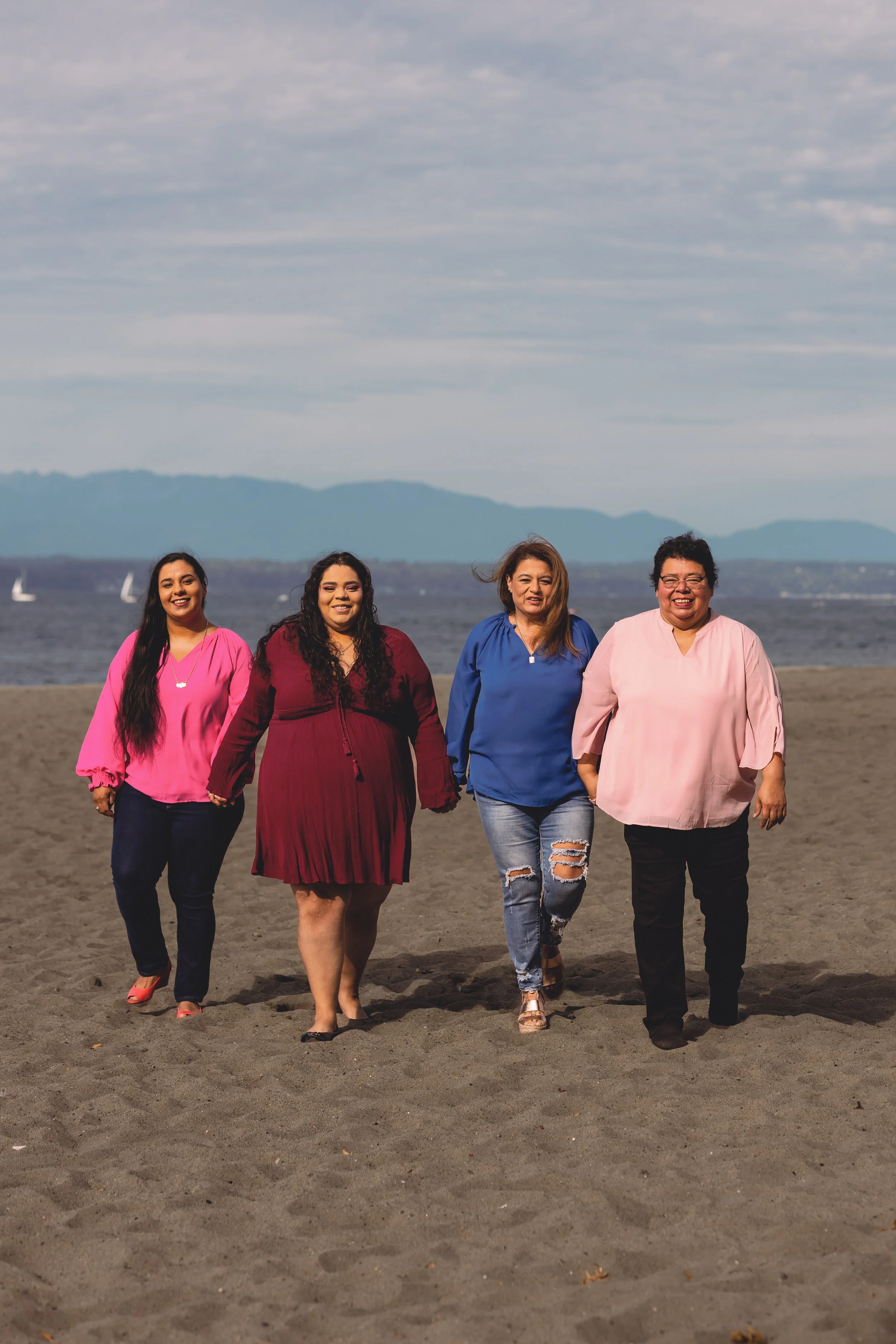 Four women walking on a sandy beach with water and sailboats in the background, smiling and holding hands.