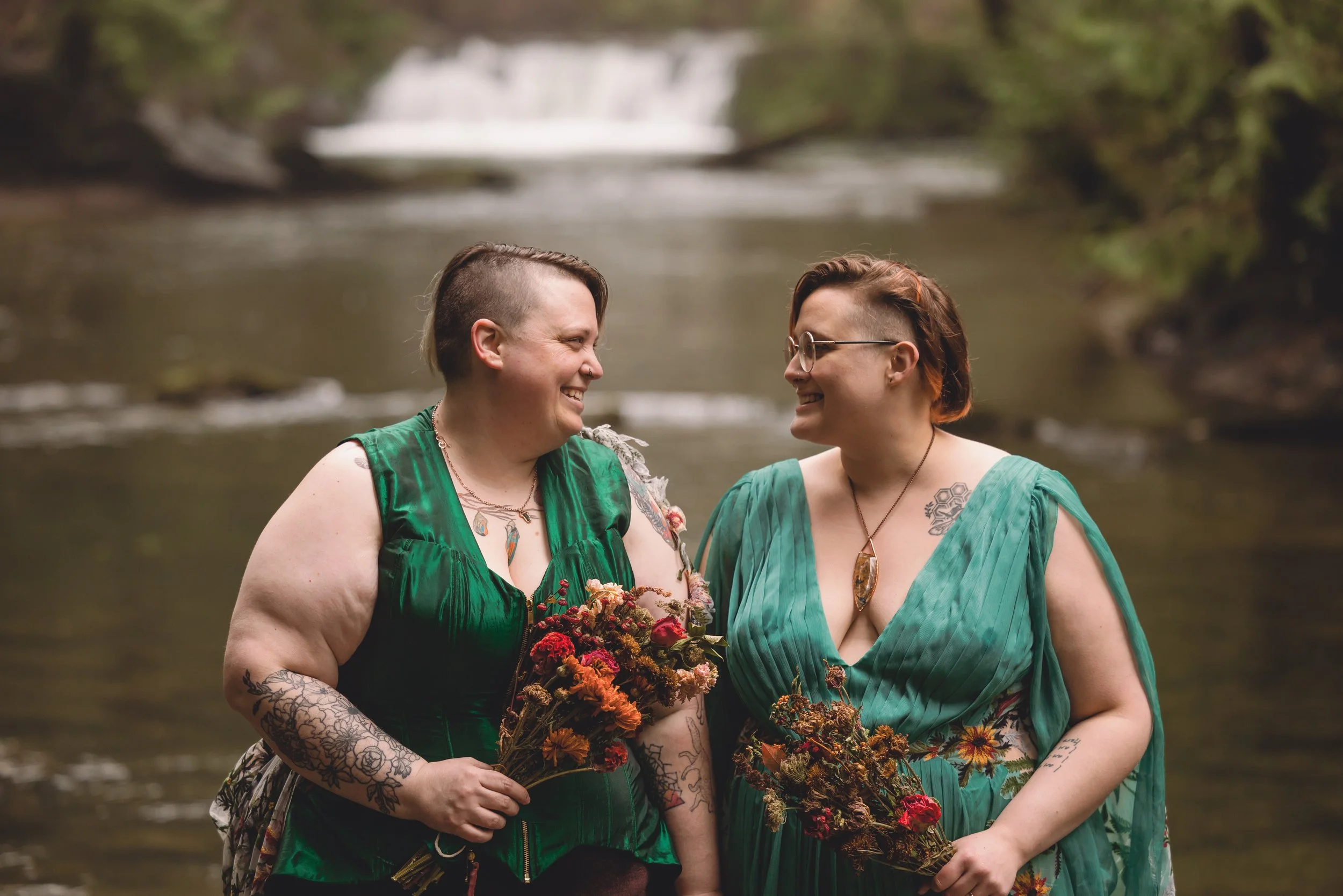 Two women in green dresses smiling at each other while holding bouquets near a river with a small waterfall in the background.