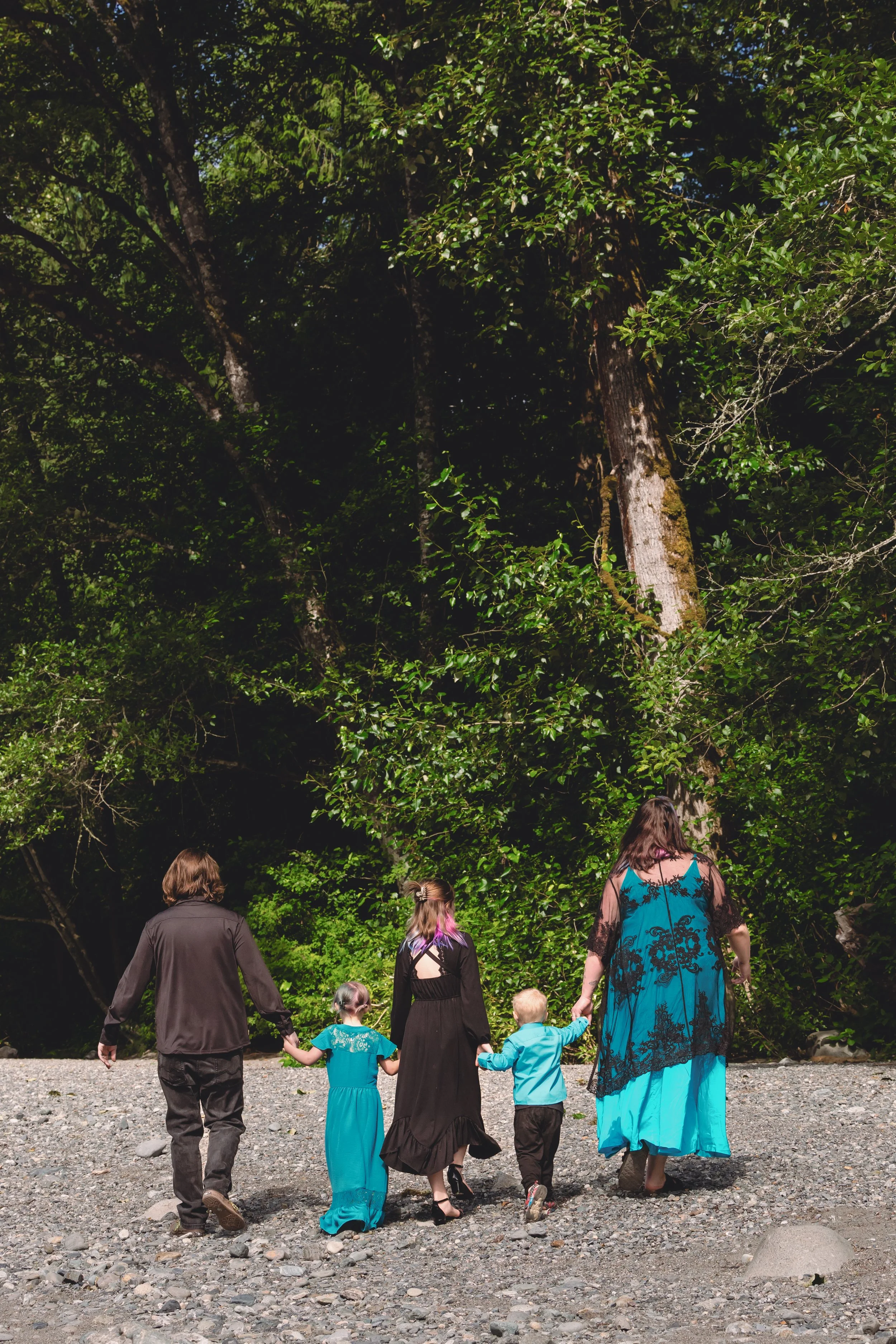 A group of five people, three children and two adults, walking together in a forested area on a gravel path. They are holding hands and facing away from the camera, surrounded by tall trees and dense foliage.