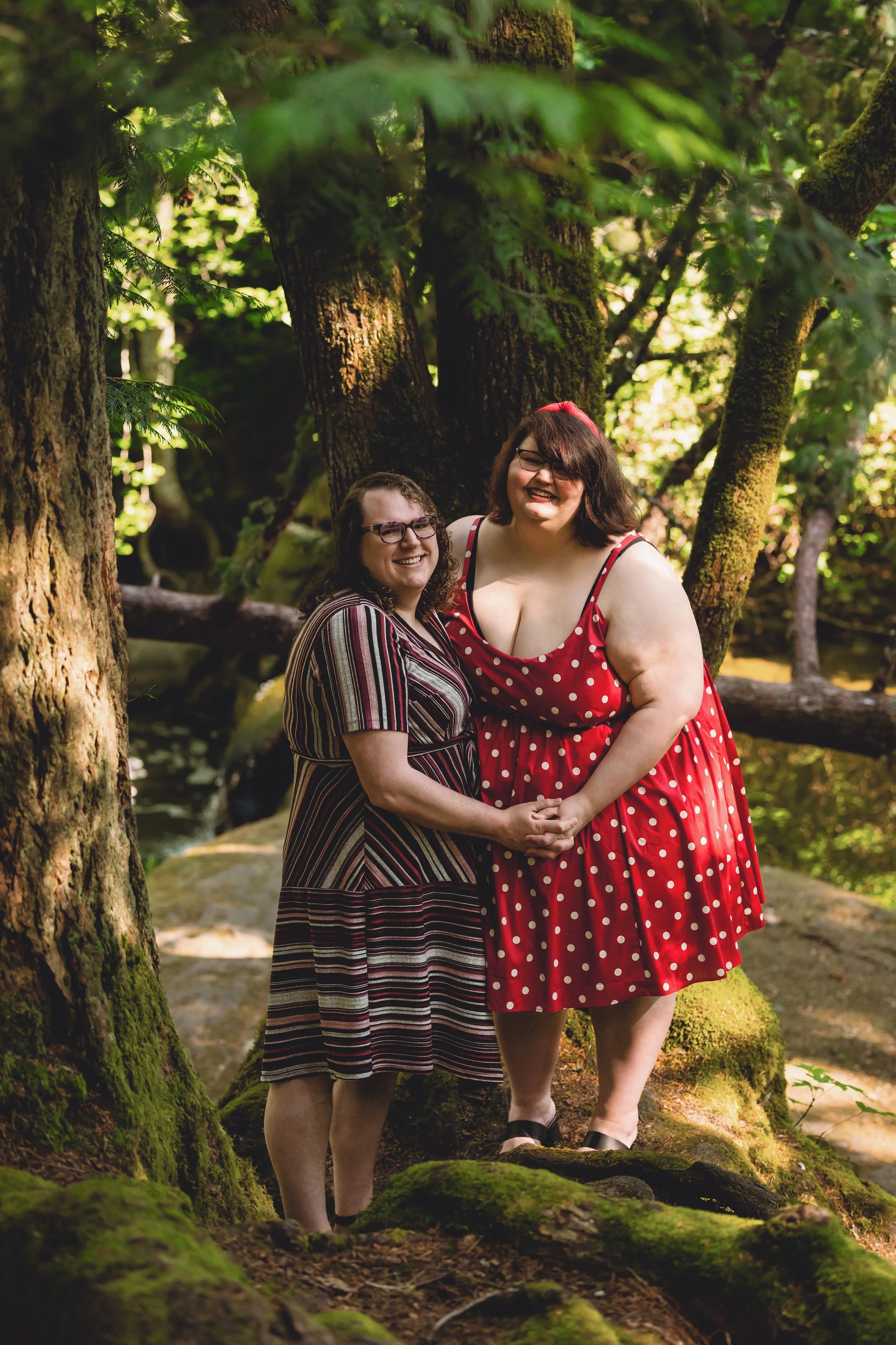 Two women holding hands and smiling in a forest surrounded by tall trees and moss-covered rocks.
