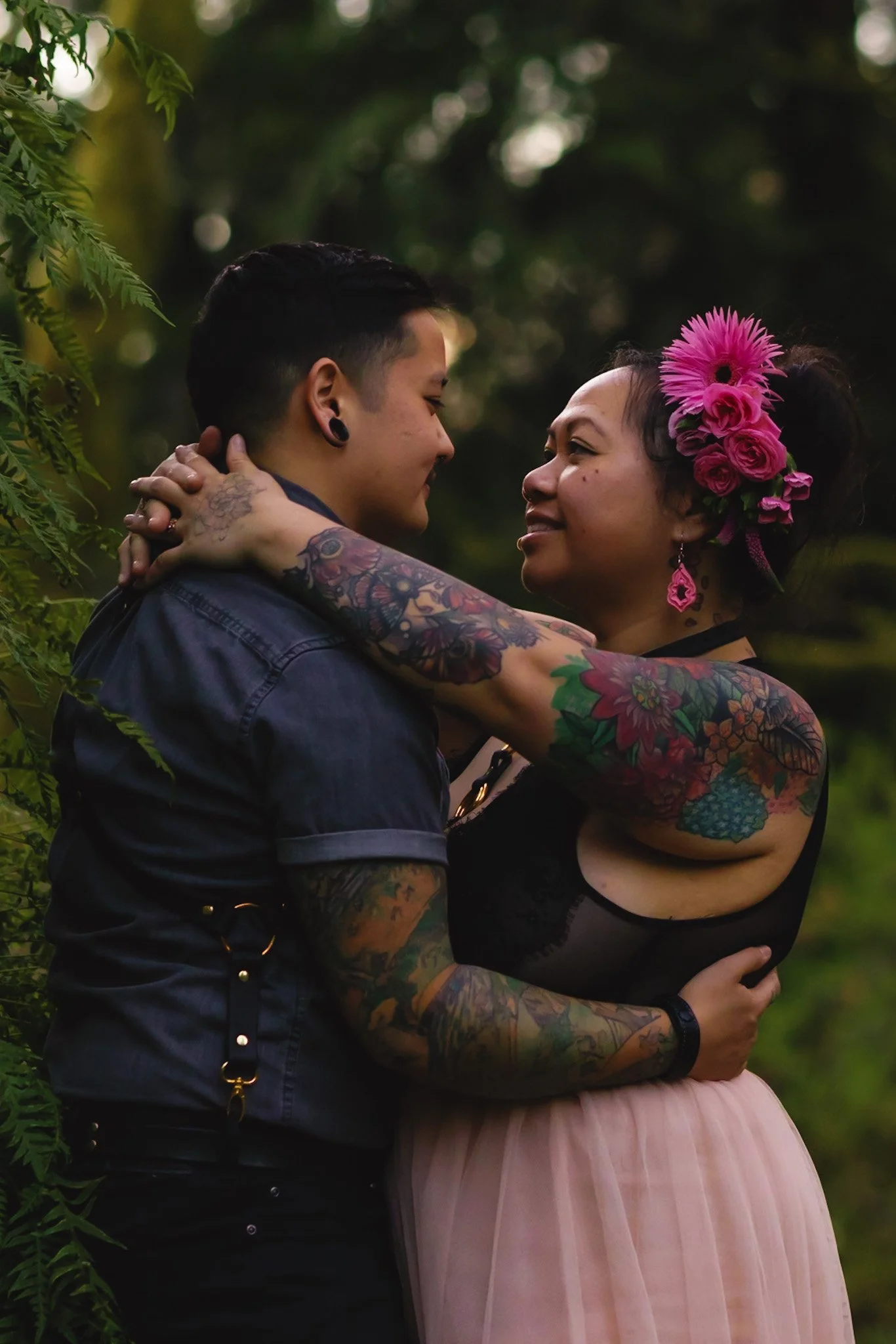 A couple with tattoos embracing in a lush, green outdoor setting, gazing into each other's eyes, with the woman wearing pink flowers in her hair and pink earrings.