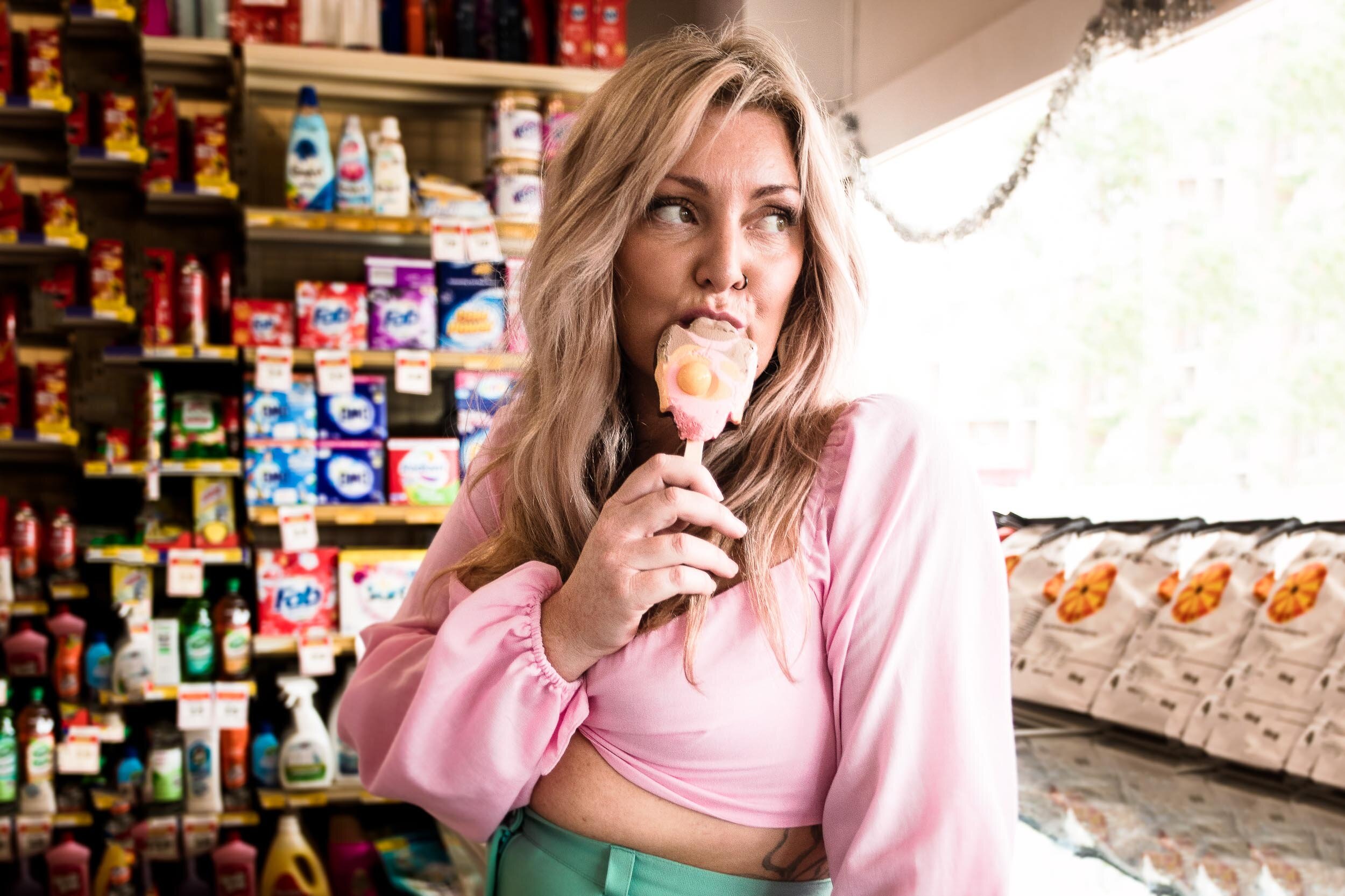 Woman with long blonde hair wearing a pink top, holding an ice cream bar in a store aisle with detergent and cleaning products.