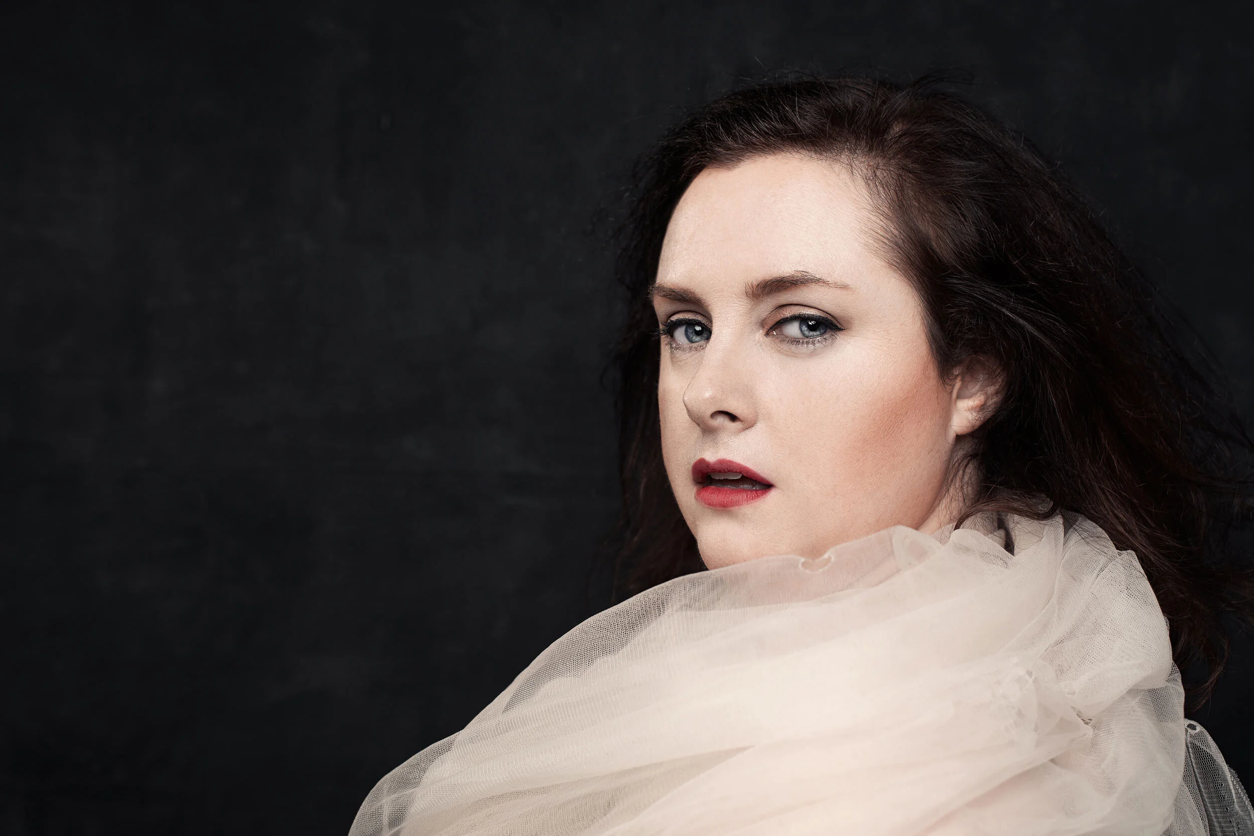 Woman with dark hair and red lipstick, wearing a sheer white fabric, against a dark background.
