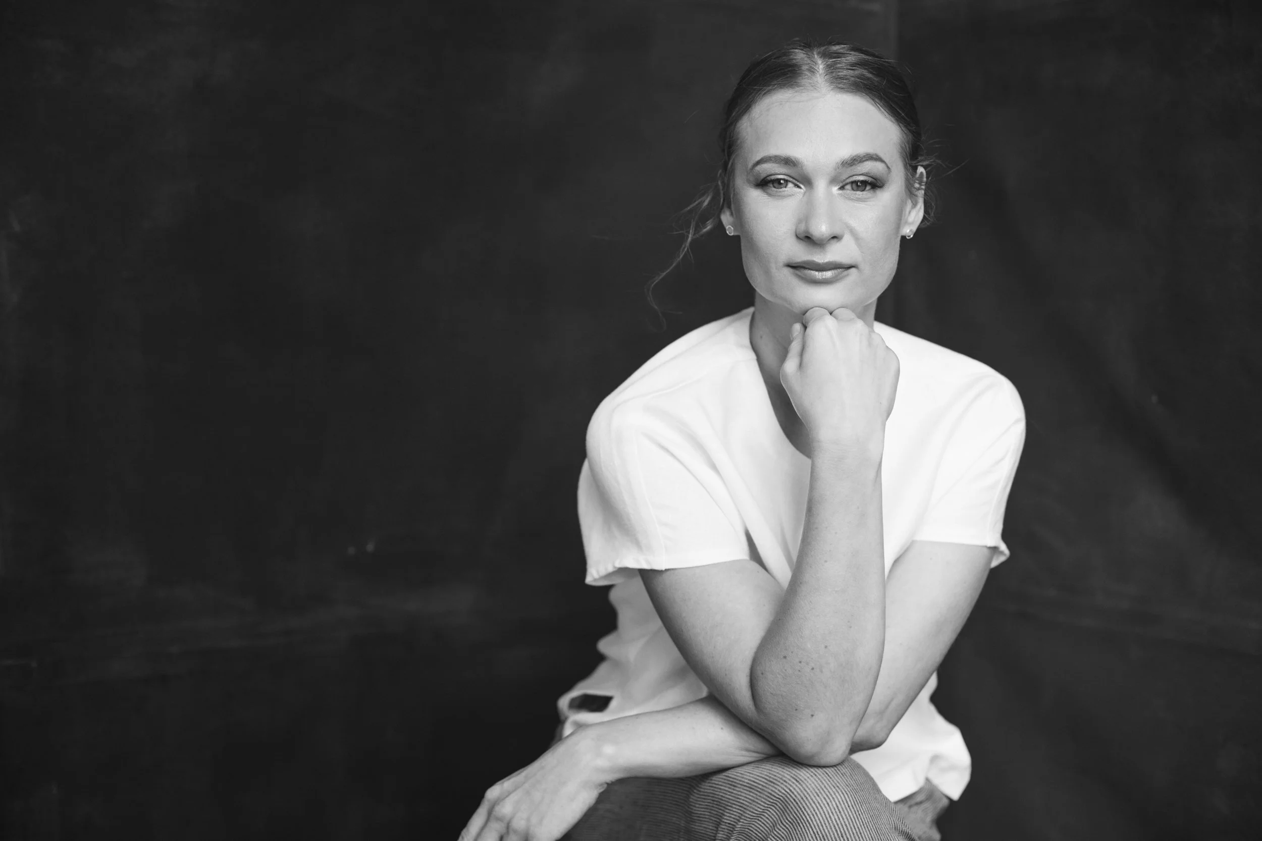Black and white portrait of a person with a thoughtful expression, wearing a white shirt, resting chin on hand, against a dark background.