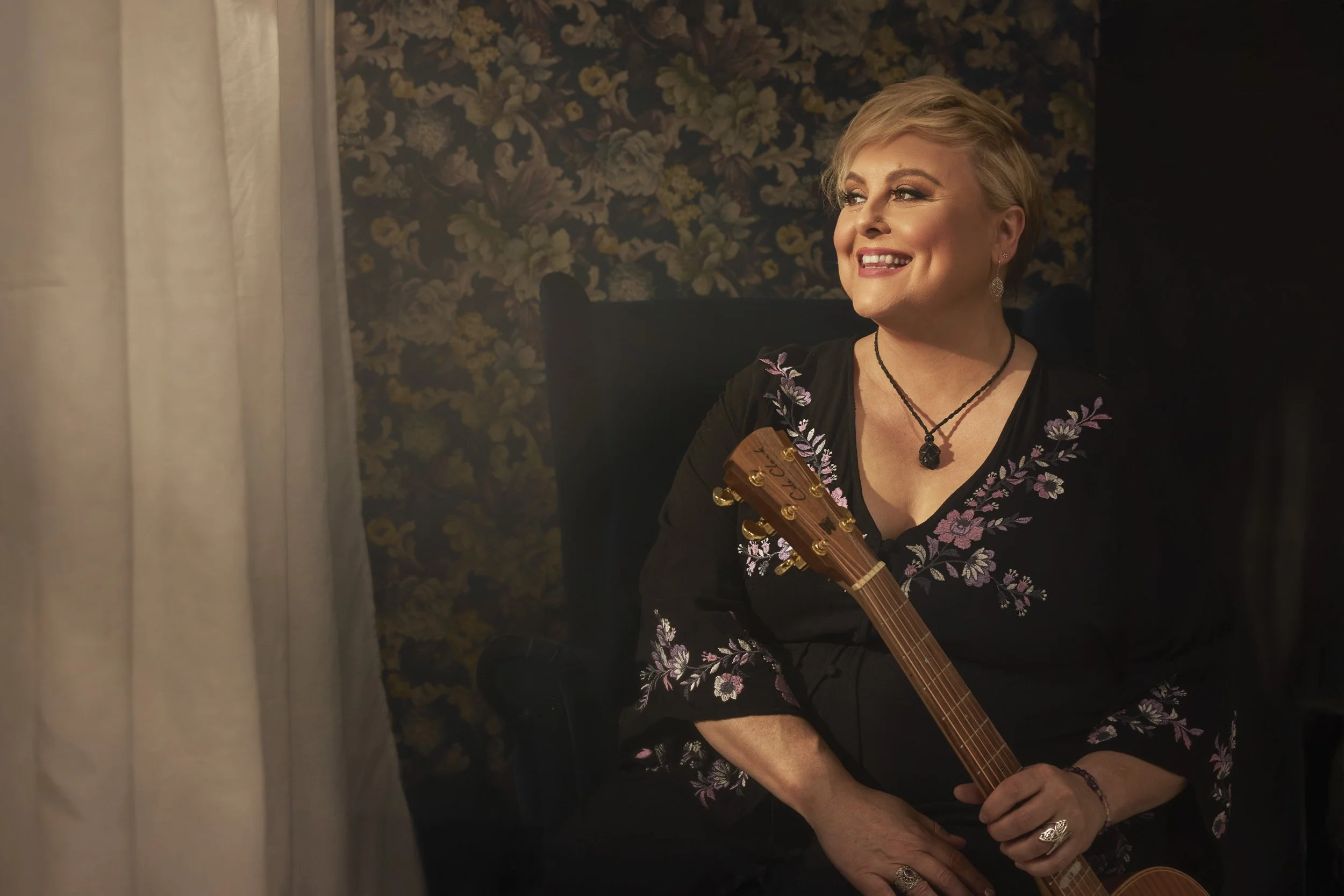 Lyn Bowtell smiling, wearing embroidered black top, holding a guitar, sitting against floral wallpaper.