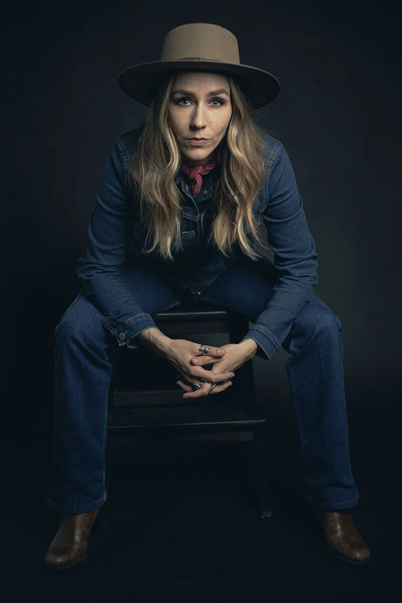 Person wearing a hat, denim jacket, and jeans, seated, in a dramatic pose, against a dark background.