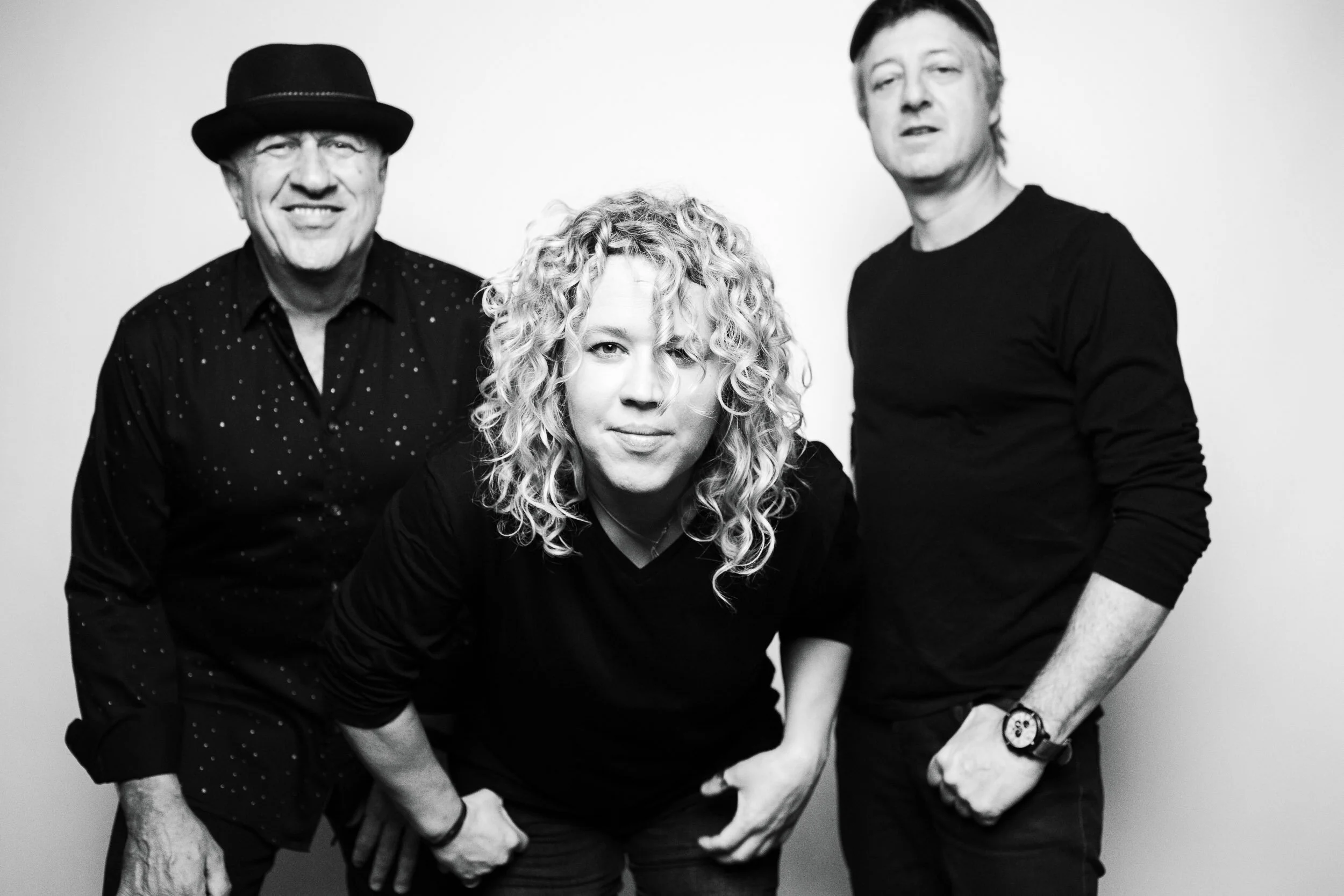 Black and white photo of three people posing; two men and a woman with curly hair in front.