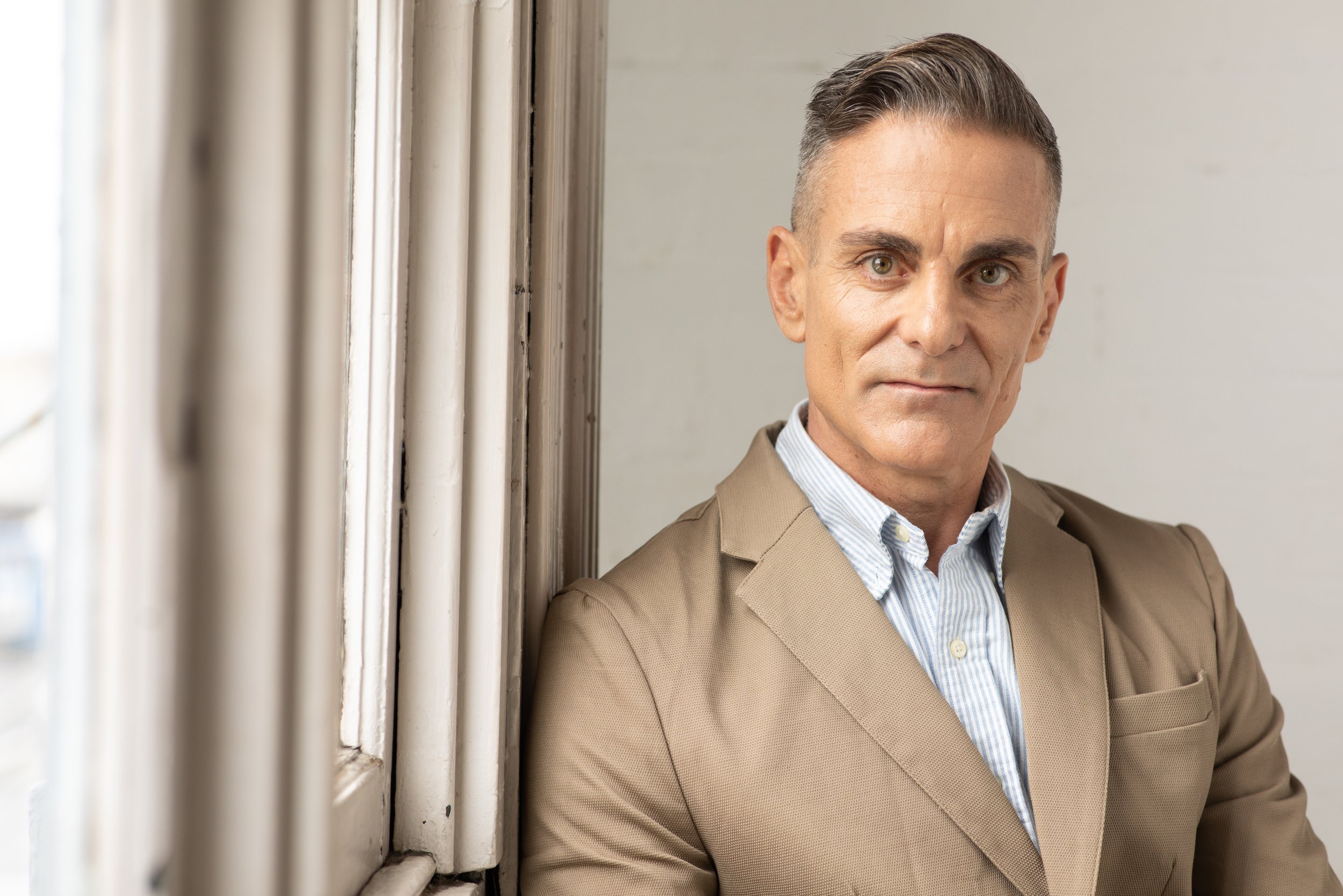 Man in a tan blazer and white shirt standing by a window, looking at the camera.