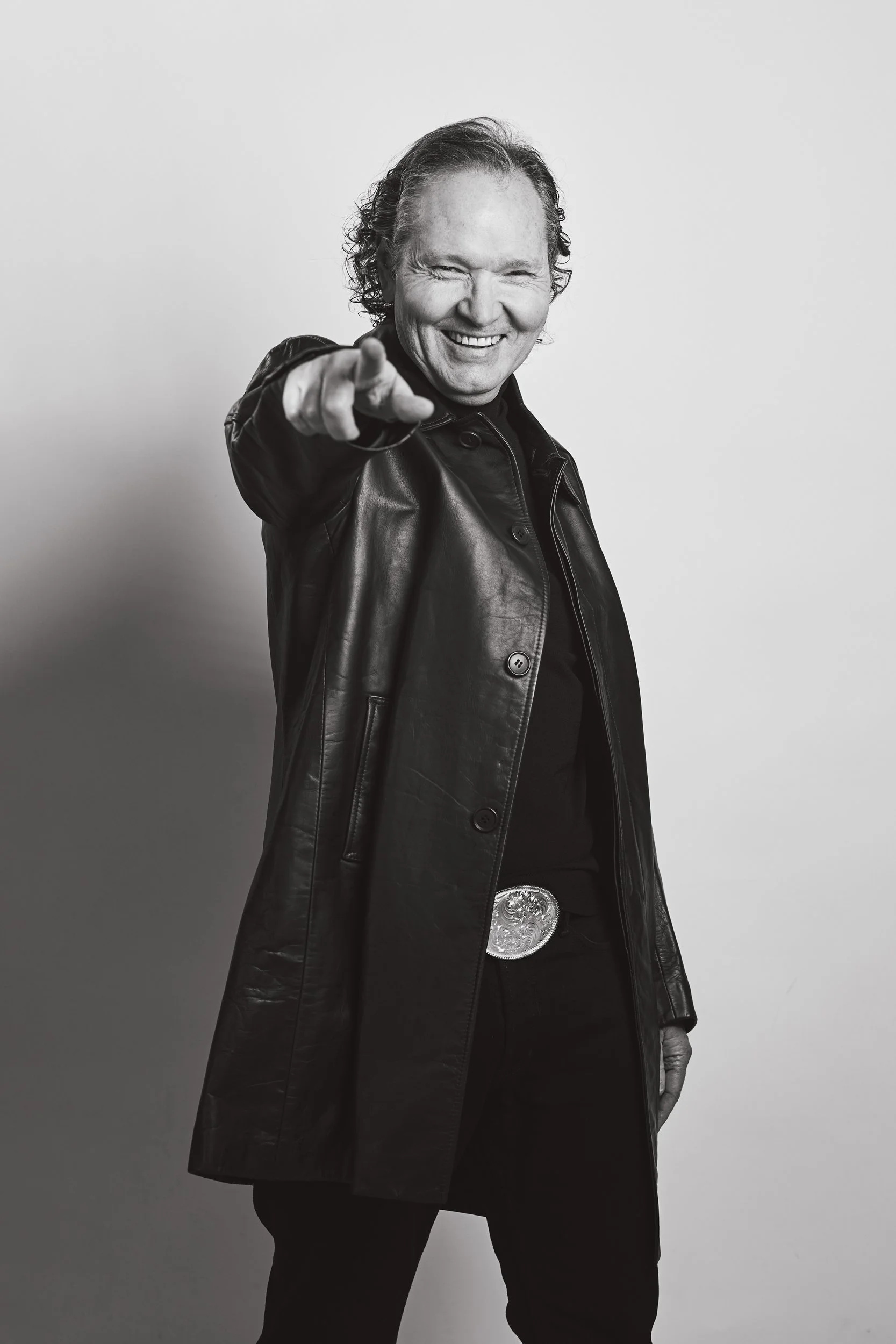 Black and white photo of a man pointing at the camera, wearing a leather jacket and a large belt buckle.