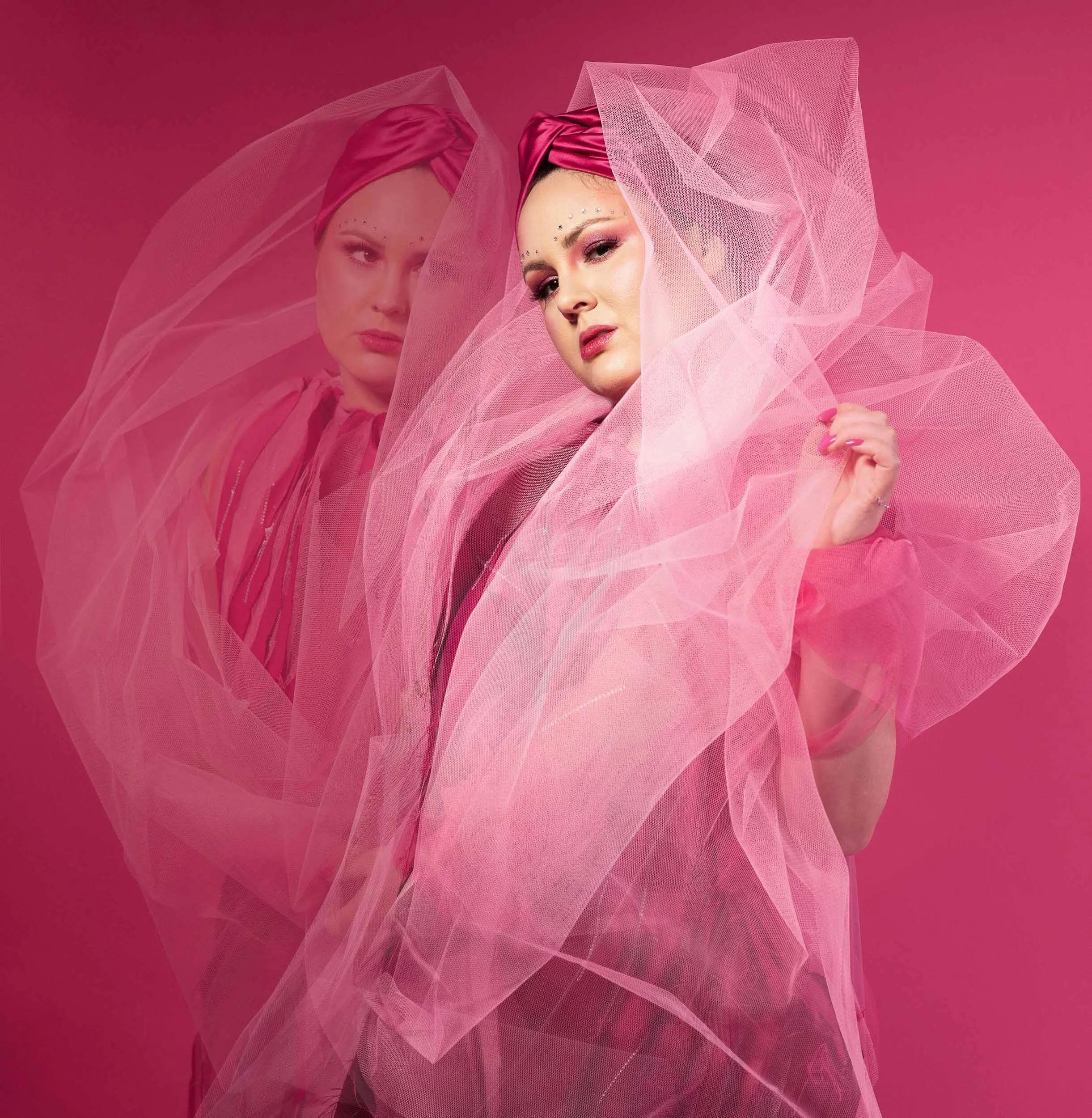Katie Brianna wearing a pink headwrap and sheer fabric, standing against a pink background with a reflection.