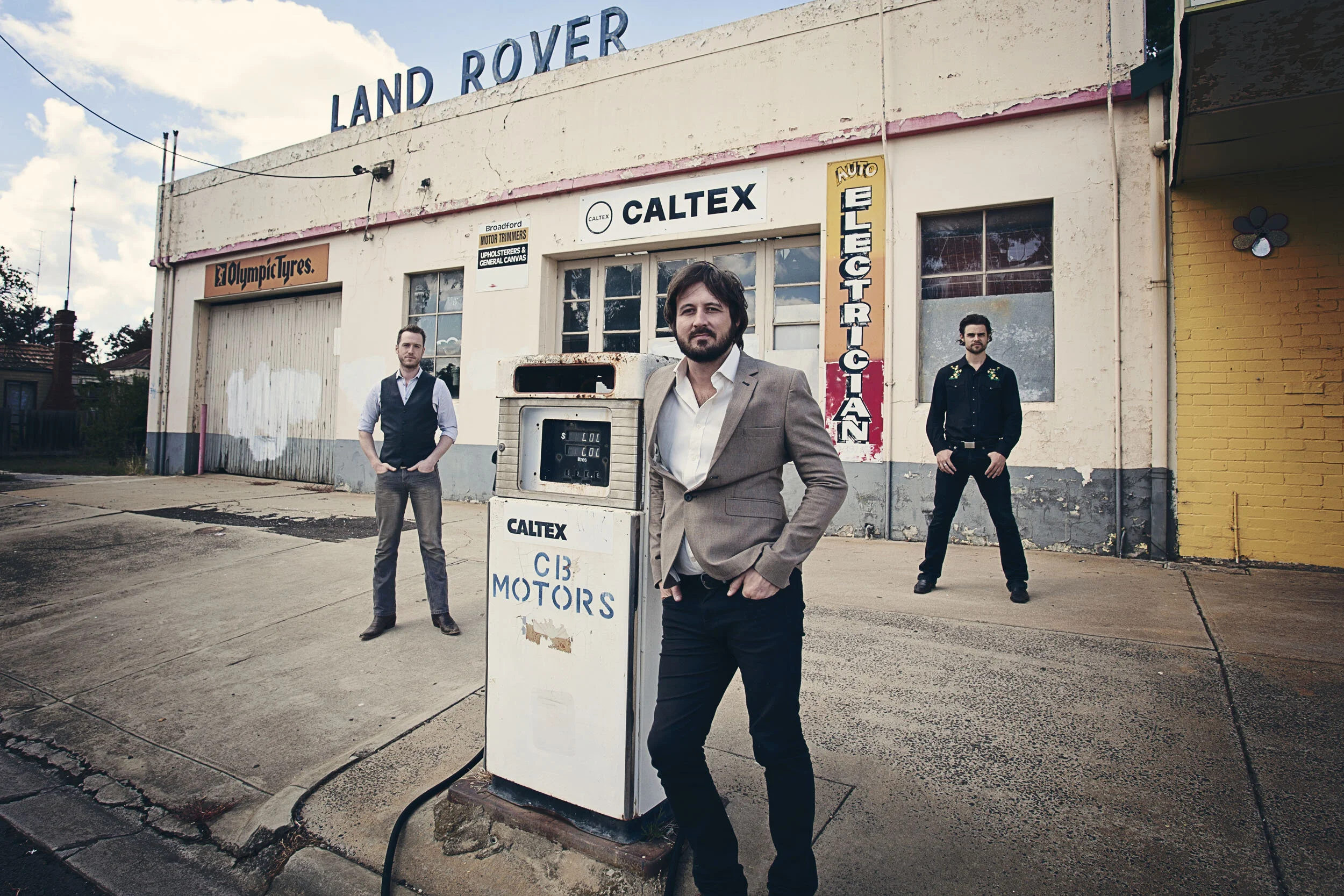 Three men standing outside an old petrol station with a vintage Caltex pump and signs, including 'Land Rover' and 'Electrician'. The building has a worn appearance.