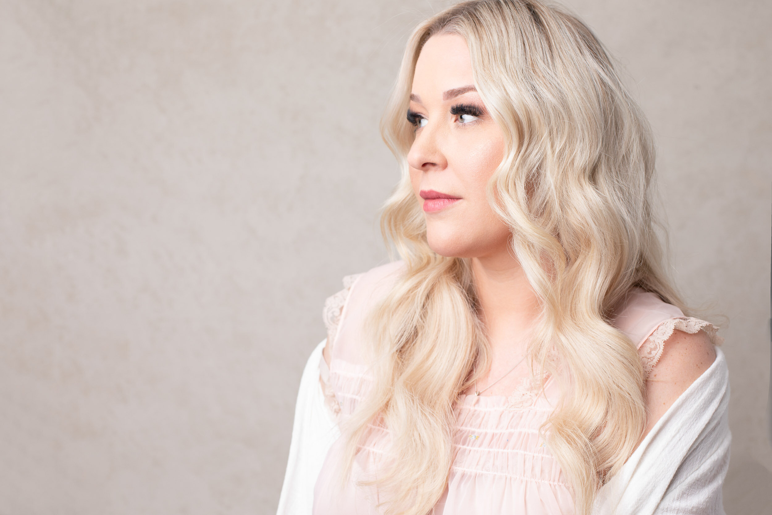 Catherine Britt with long blonde hair and soft makeup, wearing a pale pink top, looking to the side against a neutral background.