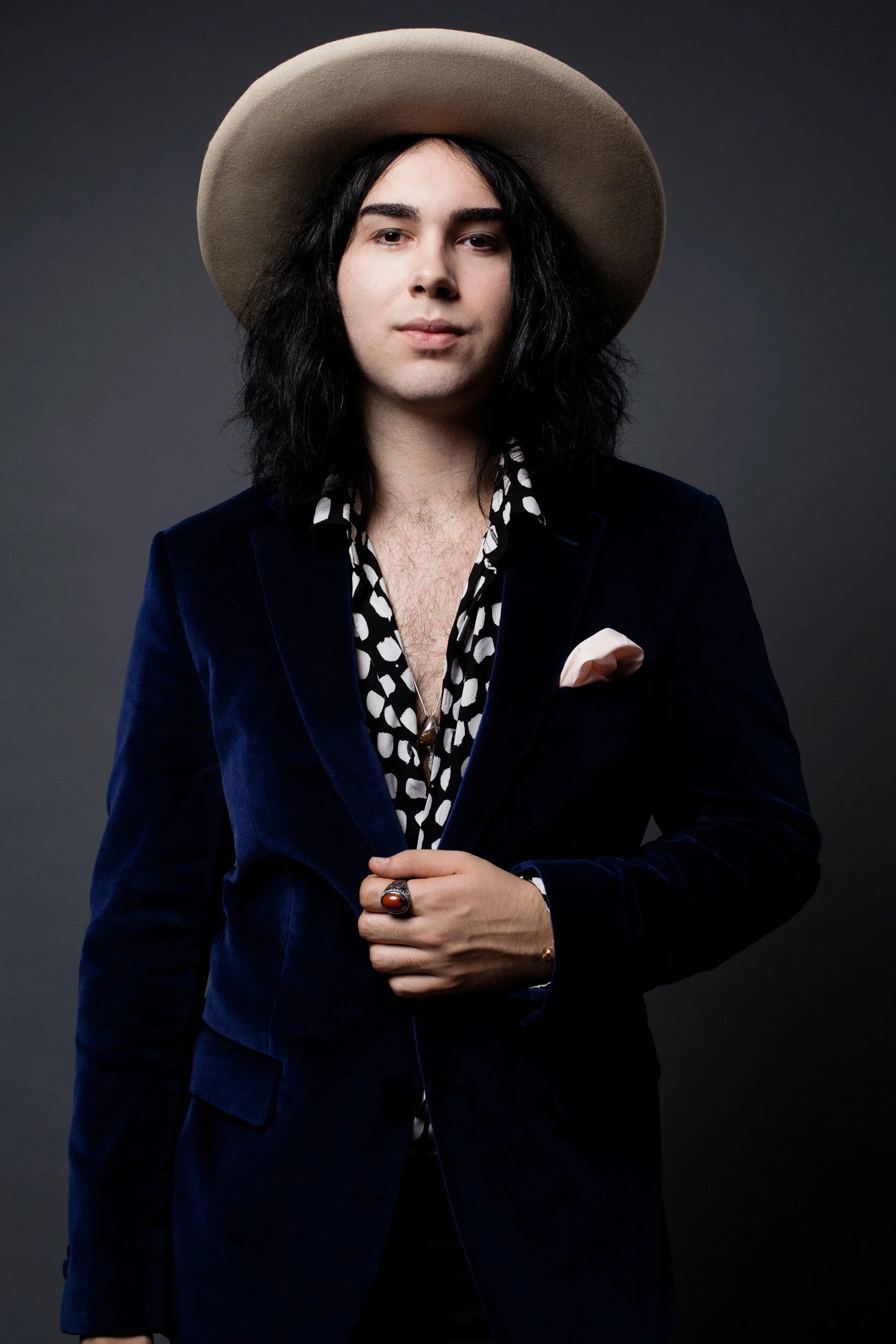 Person wearing a wide-brimmed hat, black and white patterned shirt, dark velvet jacket, and holding a ring on one finger.