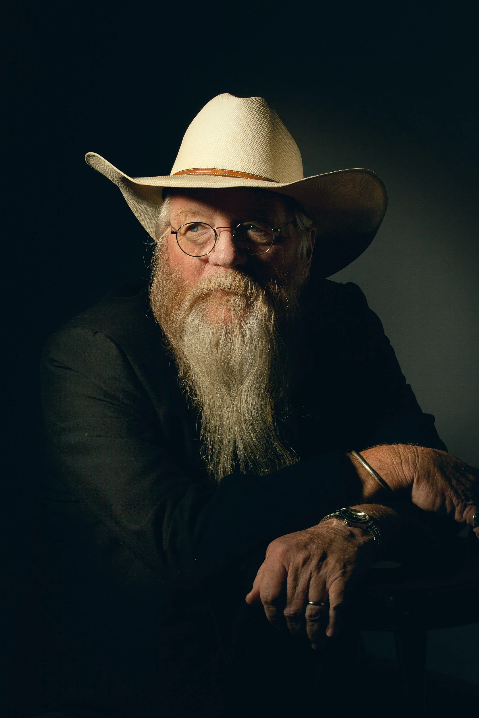 Allan Caswell with a long white beard wearing a cowboy hat and glasses in a dimly lit setting.